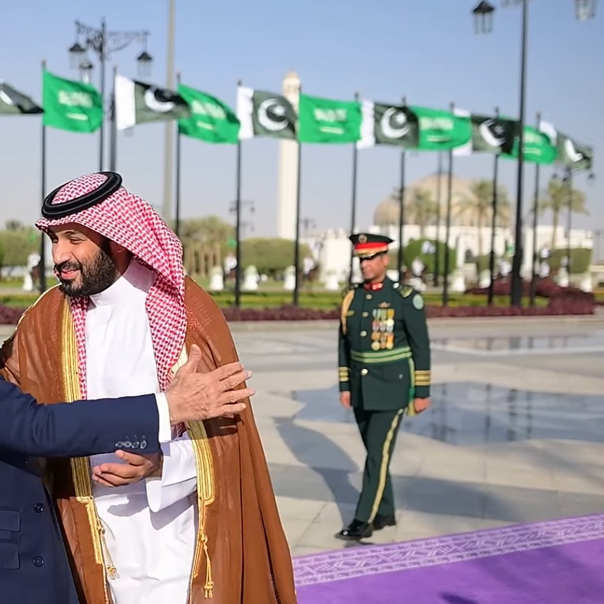 A formal greeting between two leaders, with guards and flags in the background.