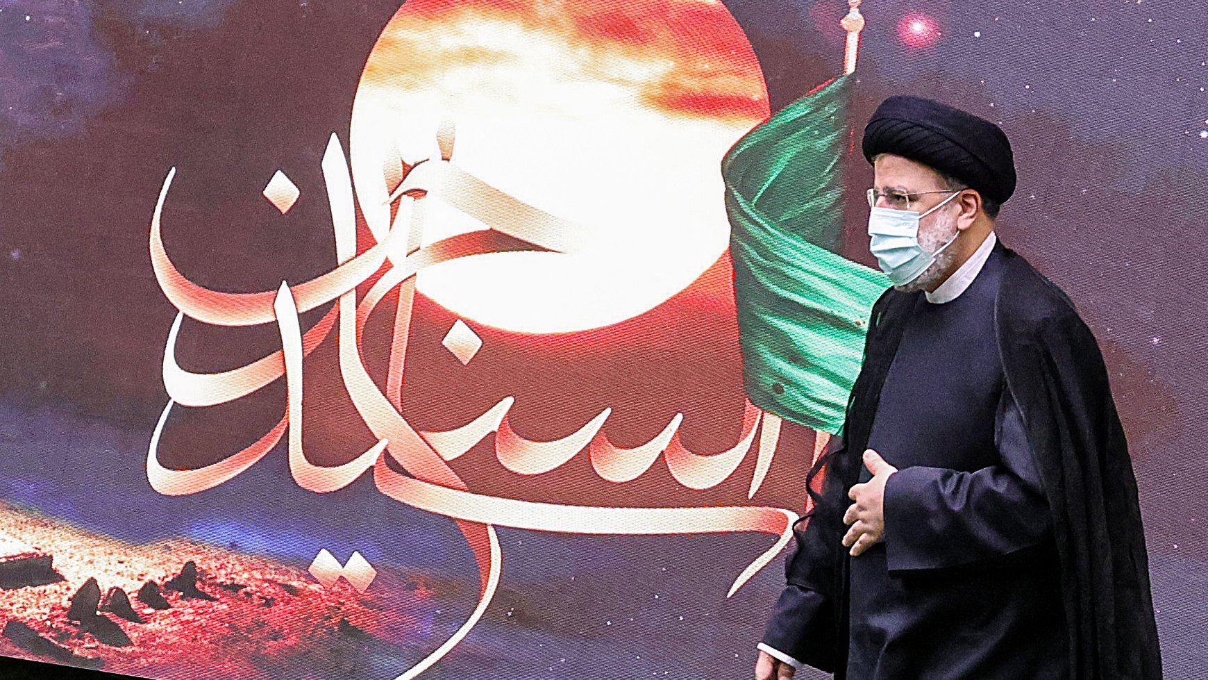 La imagen muestra a un hombre vestido con un atuendo tradicional islámico, que incluye un turbante negro y una túnica oscura. Está usando una mascarilla facial. Detrás de él, se puede ver un fondo colorido con un diseño que incluye una luna y una bandera, junto con un texto en árabe. La atmósfera parece estar relacionada con un evento significativo cultural o religioso.