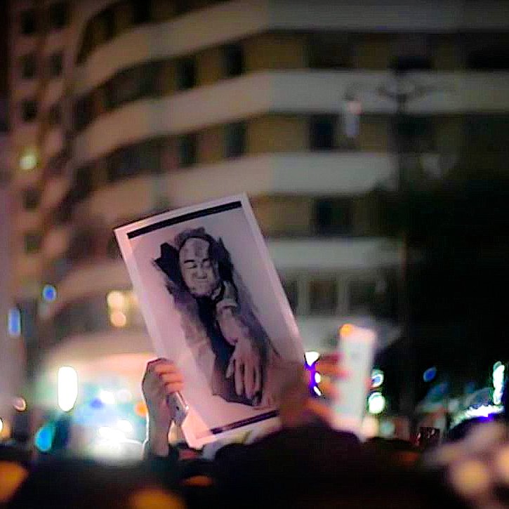 L'image montre une foule rassemblée lors d'une manifestation nocturne. Parmi les participants, une personne tient une grande affiche illustrant un portrait. Le fond de la scène est entouré de bâtiments éclairés, contribuant à l'atmosphère vibrante de l'événement. La photo semble capturer un moment important d'expression collective et de solidarité.