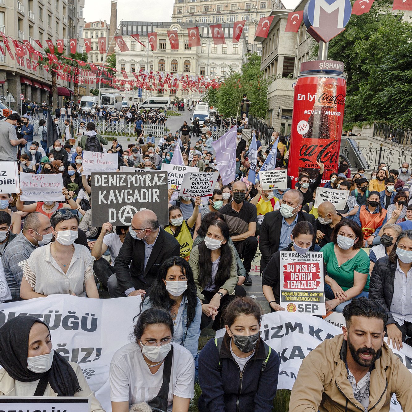La imagen muestra una manifestación en una plaza urbana, con varias personas sentadas en el suelo. Los participantes sostienen pancartas y carteles que tienen mensajes de protesta. La mayoría de las personas llevan mascarillas, lo que puede indicar que la manifestación está ocurriendo en un contexto reciente de pandemia. Al fondo, se pueden ver banderas turcas y edificios, lo que sugiere que la protesta tiene lugar en Turquía. La atmósfera es de movilización y reclamo social.