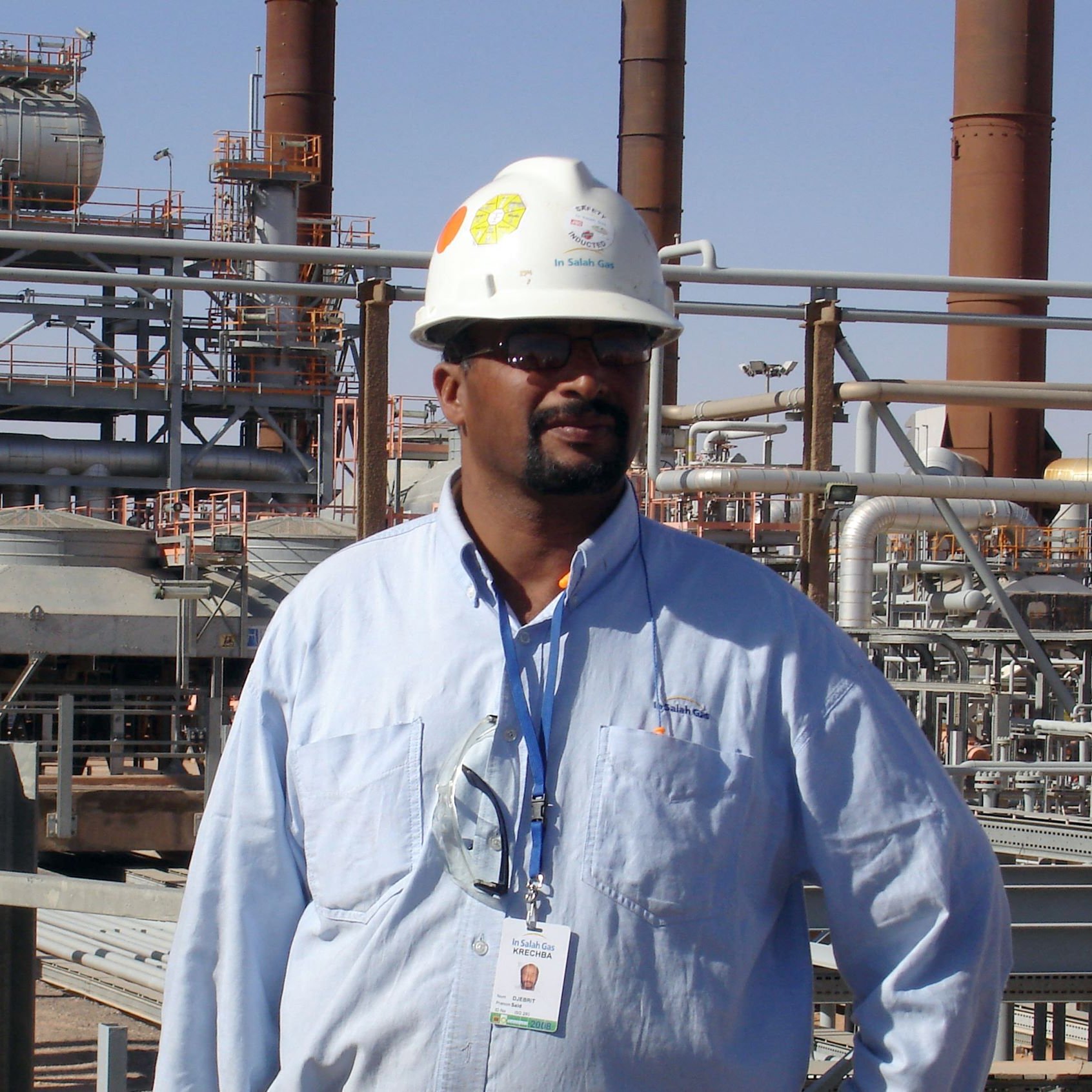 L'image montre un homme portant un casque de sécurité blanc et une chemise bleue, debout dans un environnement industriel. En arrière-plan, on aperçoit des structures métalliques et des installations, probablement liées à une usine ou une raffinerie. L'homme semble professionnel et attentif, avec une carte d'identification suspendue à son cou.
