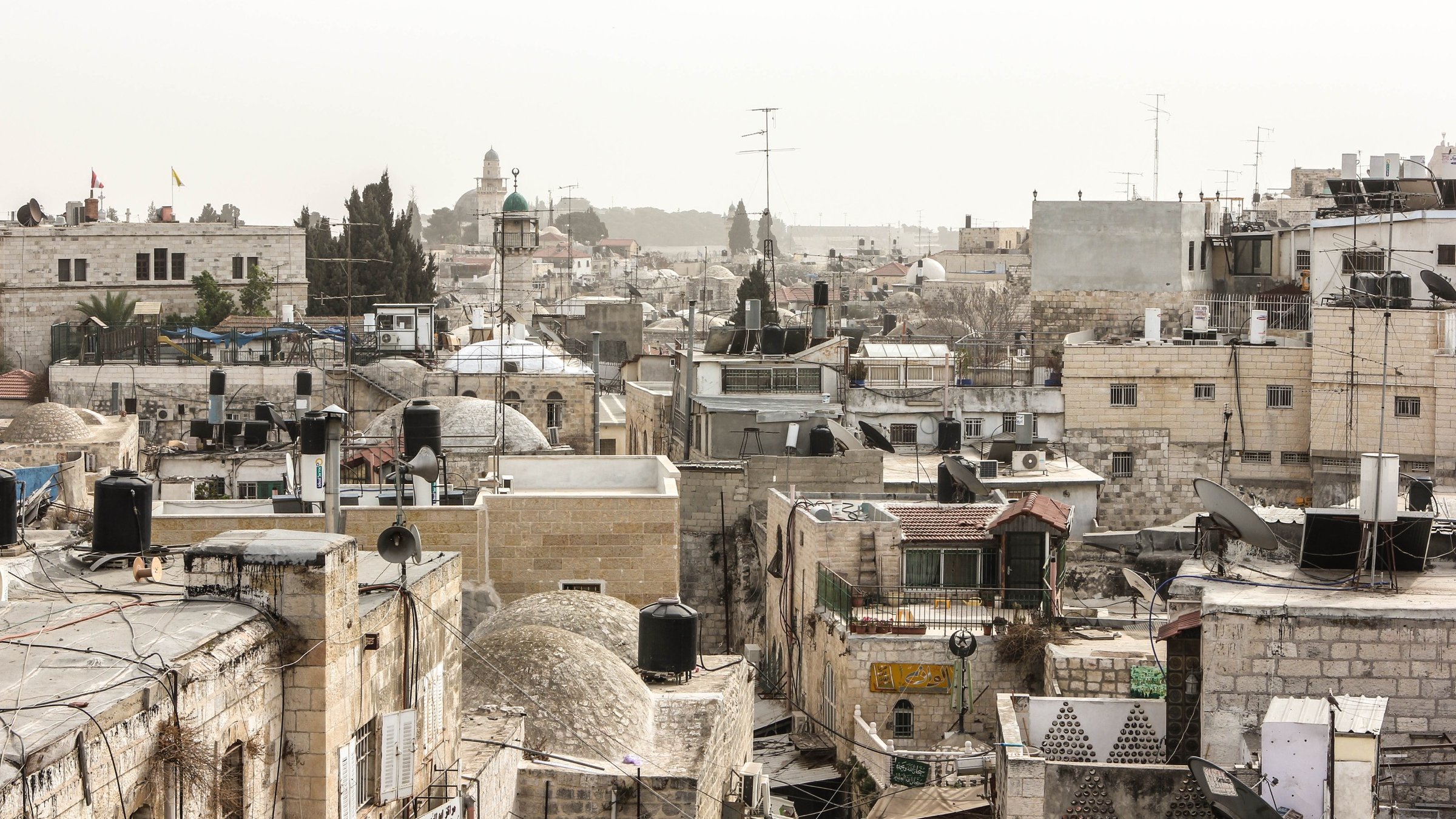 الصورة تُظهر منظرًا لأحد الأحياء القديمة، حيث تتداخل المباني التقليدية مع الأسطح المسطحة وأجهزة التكييف. يوجد أيضًا بعض الأشجار ومآذن المساجد في الخلفية، مما يشير إلى طابع تاريخي وديني للمنطقة. السماء تبدو مائلة إلى الرمادي، مما قد يدل على أجواء غائمة أو ضبابية.