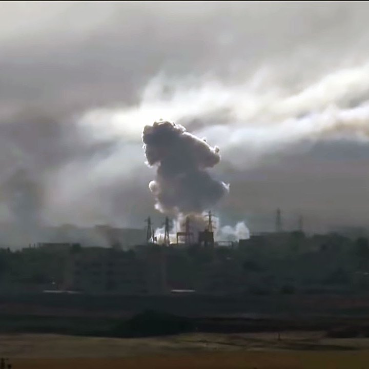 La imagen muestra una escena con humo denso y nubes oscuras que se elevan desde un área urbana. Se aprecian instalaciones industriales o edificios en el fondo, y el humo parece provenir de una explosión o un incendio. El cielo está nublado, lo que contribuye a una atmósfera sombría y tensa en la imagen.