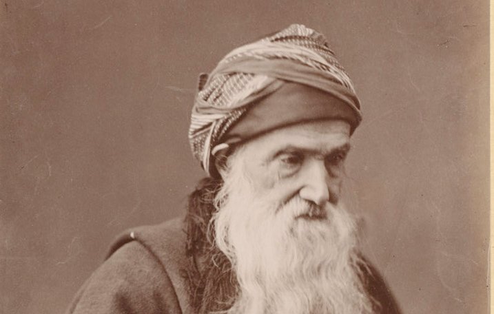 L'image montre un homme âgé avec une longue barbe blanche. Il porte un turban ou une coiffe enroulée autour de la tête, ainsi qu'une cape sombre. Son regard est concentré et il tient une canne. L'arrière-plan est neutre, mettant en valeur son visage et son expression, ce qui donne une impression de sagesse et de dignité.