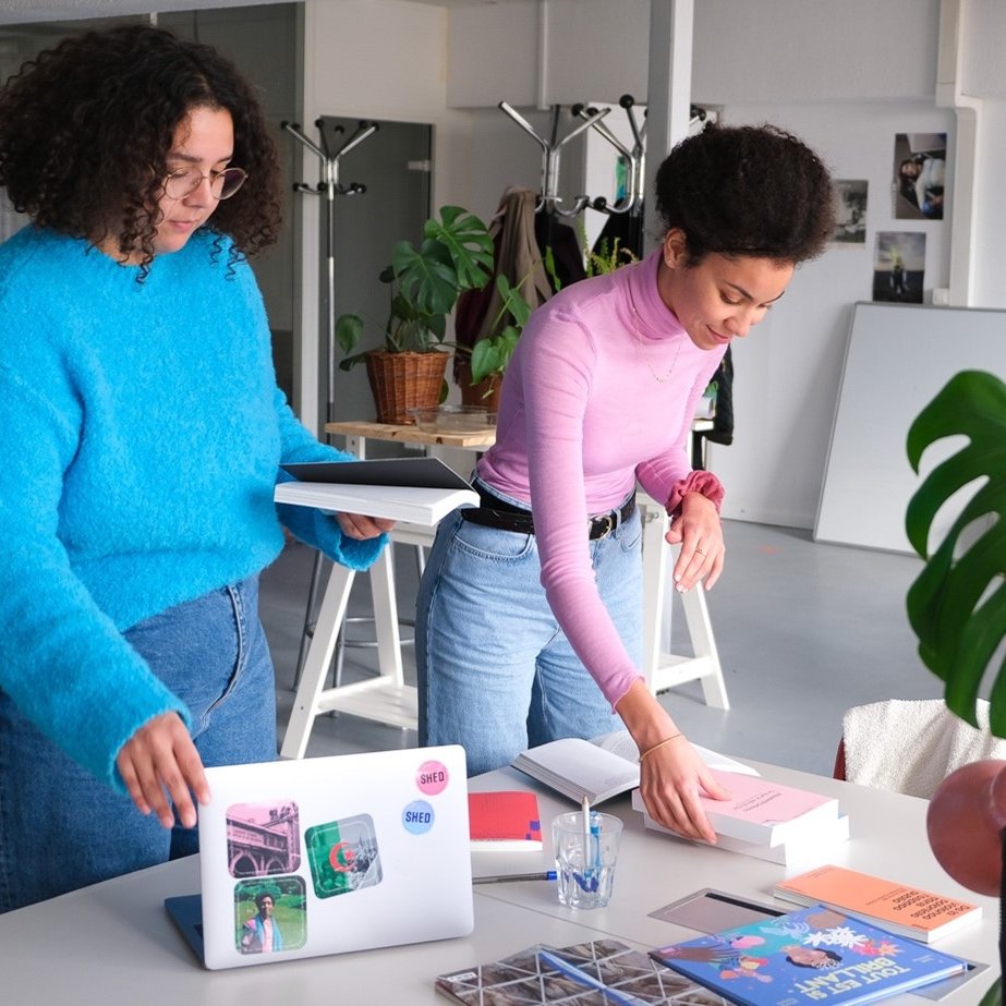 في الصورة، تظهر امرأتان يعملان معًا في مكتب عصري. إحدى السيدتين ترتدي سويتر زرقاء ومرفع، بينما الأخرى ترتدي سويتر وردي وبنطلون جينز. يبدو أنهما مشغولتان بتنظيم بعض الملفات أو الكتب على الطاولة. يوجد على الطاولة كمبيوتر محمول وبعض المقتنيات مثل الكوسات والكتب والمجلة. الخلفية تحتوي على نباتات وأثاث بسيط، مما يعكس جوًا من الإبداع والتعاون.