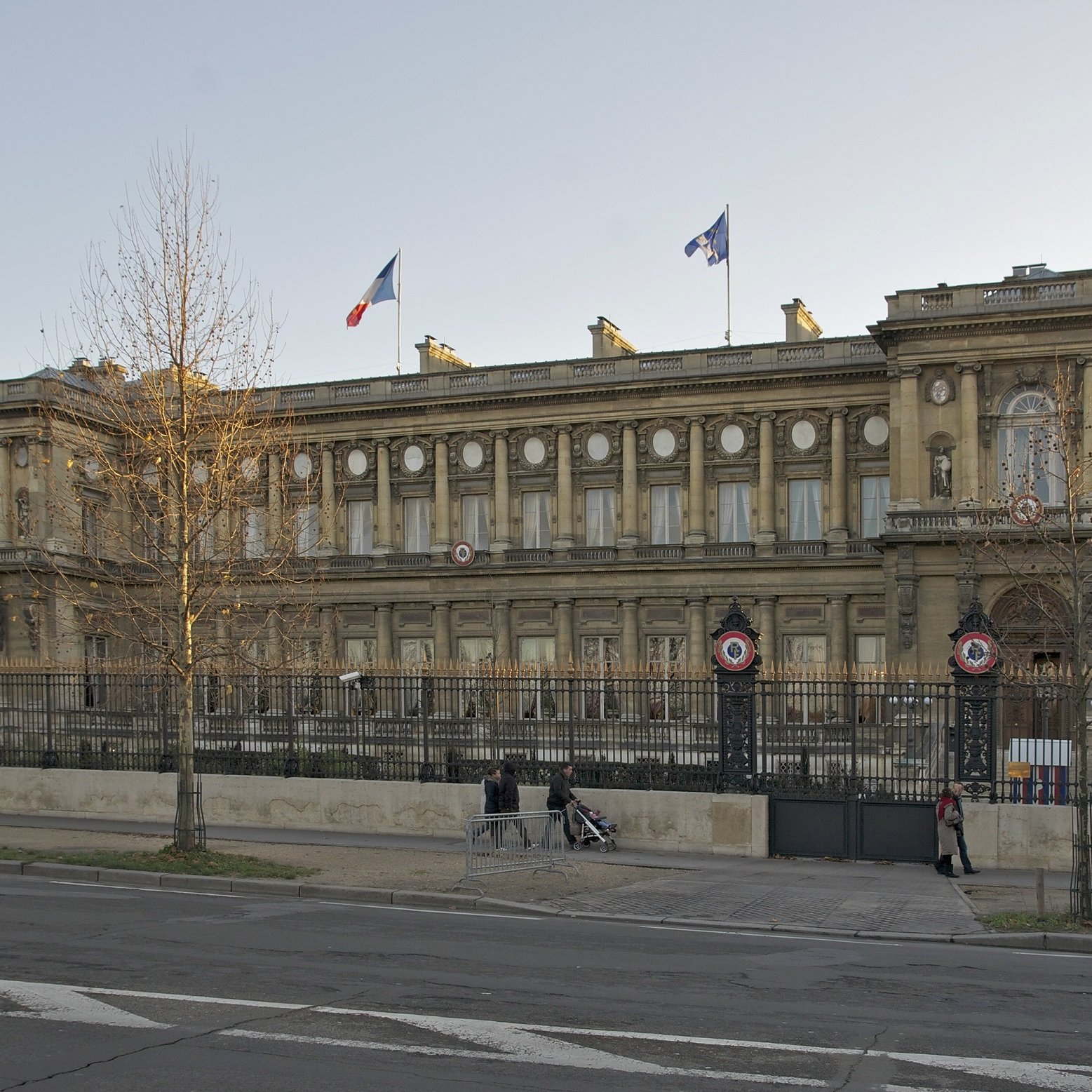 The image depicts a grand building with a classical architectural style, likely a government or official structure. The facade features ornate details, including large windows and decorative elements. French flags are visible atop the building, indicating its significance. There is a wrought iron fence in front, along with columns that enhance the structure's elegance. The surrounding area appears to be urban, with a street visible in the foreground, suggesting the building is located in a city. The overall scene conveys a sense of history and importance.