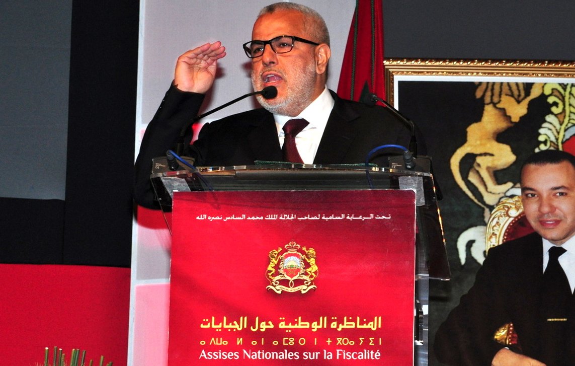 الصورة تظهر شخصًا معروفًا يتحدث على منصة، حيث يبدو أنه يلقي خطابًا رسميًا. خلفه يمكن رؤية شعار رسمي أو صورة رمزية. المنصة تحمل عنوان "المناظرة الوطنية حول الأداءات"، مما يشير إلى أن الحدث يتعلق بالمالية أو الضرائب. الجو العام يبدو رسميًا وجديًا، ولغة الجسد تعكس الاهتمام والالتزام.