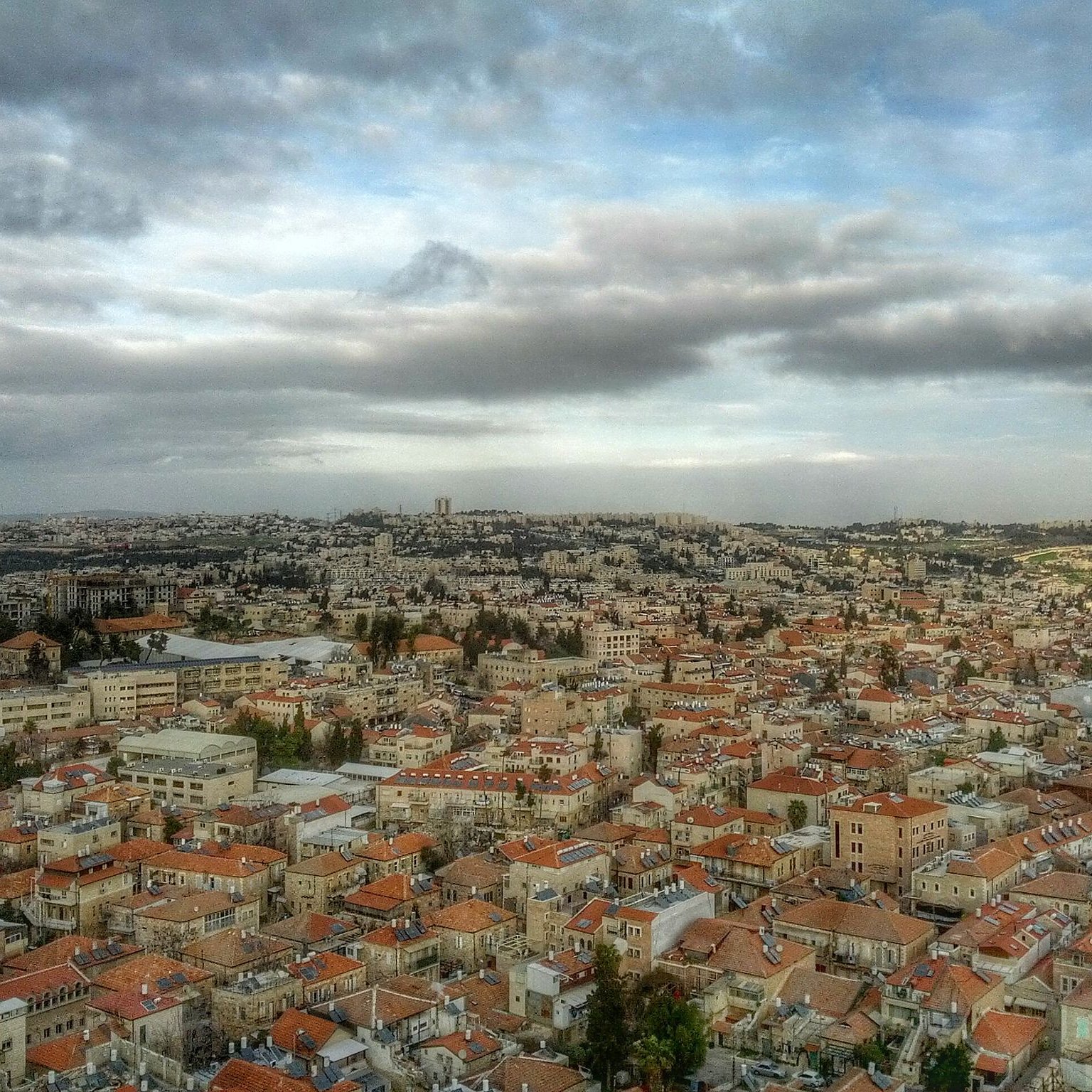 L'image montre une vue panoramique d'une ville, probablement Jérusalem, avec des bâtiments disposés sur plusieurs collines. Les toits sont principalement en tuiles rouges, et il y a de nombreux édifices en pierre. Le ciel est nuageux, ajoutant à l'ambiance de la scène, avec une lumière douce qui éclaire la ville. On peut également apercevoir des zones de verdure entre les bâtiments, ce qui ajoute de la profondeur au paysage urbain.