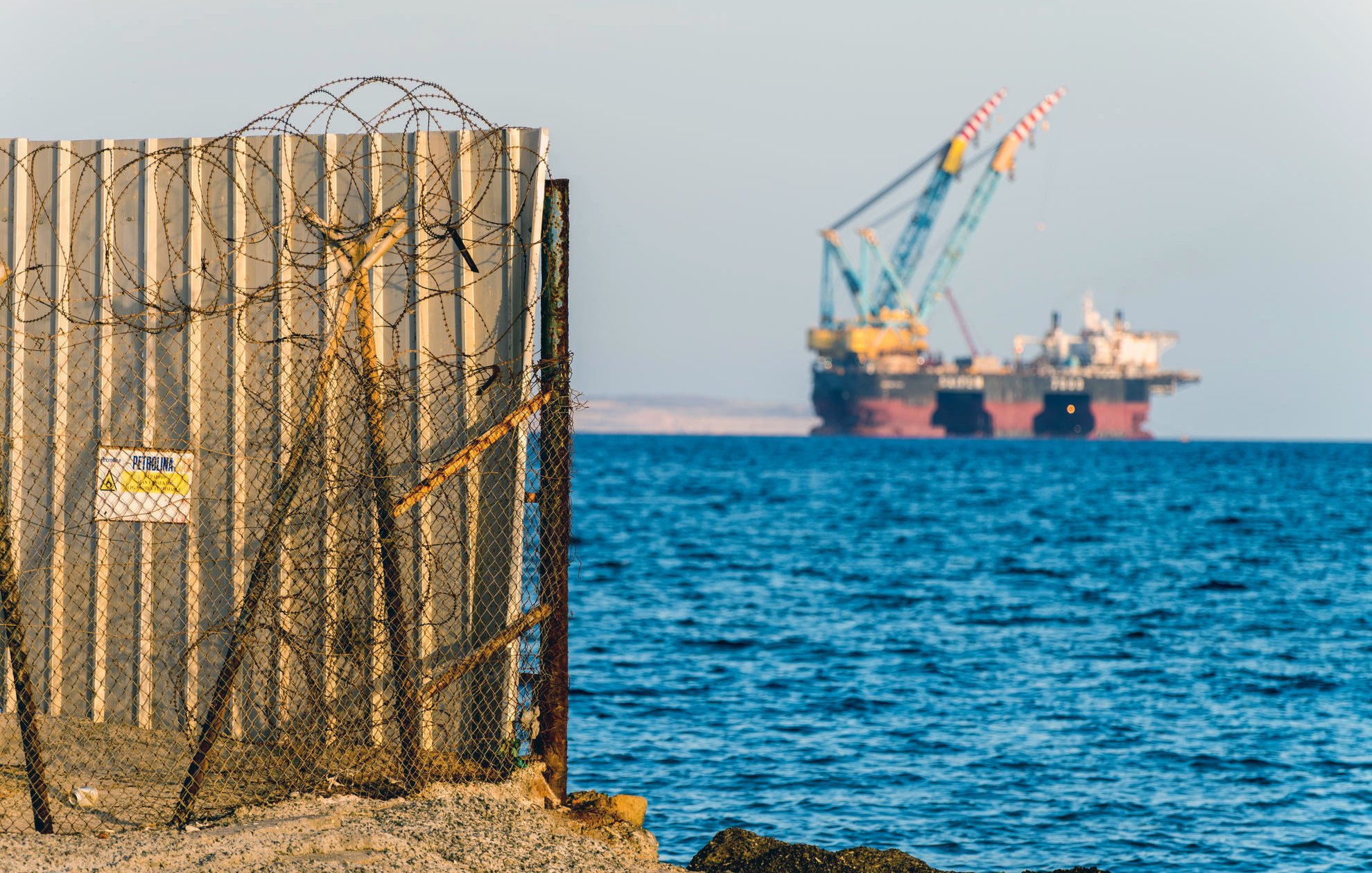 L'image montre une barrière en métal, probablement un mur de séparation, avec du fil barbelé à sa base. En arrière-plan, on aperçoit des barges ou des plateformes offshore, avec des grues, sur un fond d'eau calme. Le ciel semble dégagé, et l'ensemble donne une impression de contraste entre la structure humaine et l'environnement maritime.