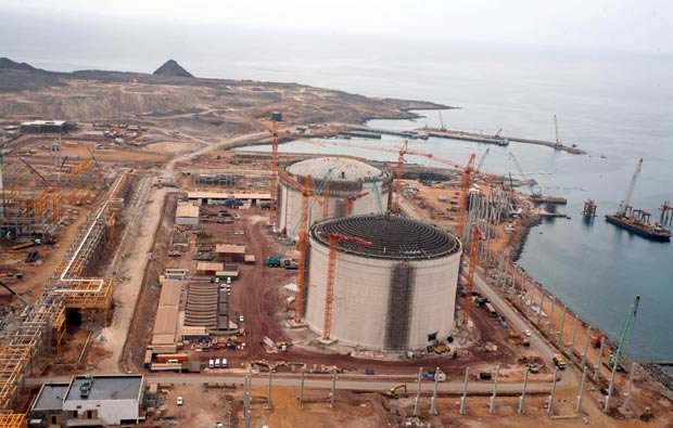 L'image montre un site industriel en construction, probablement une installation de gaz naturel ou une centrale électrique. On peut voir plusieurs grands réservoirs cylindriques, entourés de grues et de matériaux de construction. L'environnement est côtier, avec une vue sur la mer et des montagnes en arrière-plan. Il y a également des routes et des véhicules de construction présents sur le site. Le paysage est principalement désertique et montagneux.