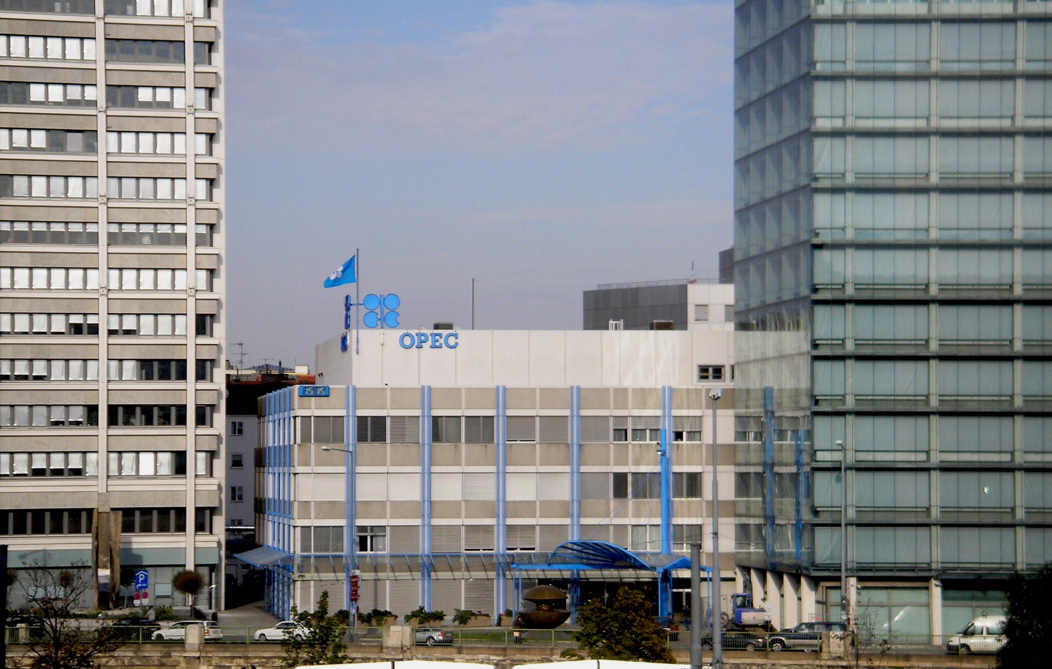 L'image montre le bâtiment de l'OPEP (Organisation des pays exportateurs de pétrole), qui est un bâtiment moderne à plusieurs étages. Des drapeaux sont visibles sur le toit, y compris celui de l'OPEP. Le bâtiment est entouré par d'autres structures urbaines, reflétant un environnement de ville. Le ciel est légèrement nuageux, suggérant une journée calme. Des éléments architecturaux en verre et en béton contrastent avec le design du bâtiment de l'OPEP.
