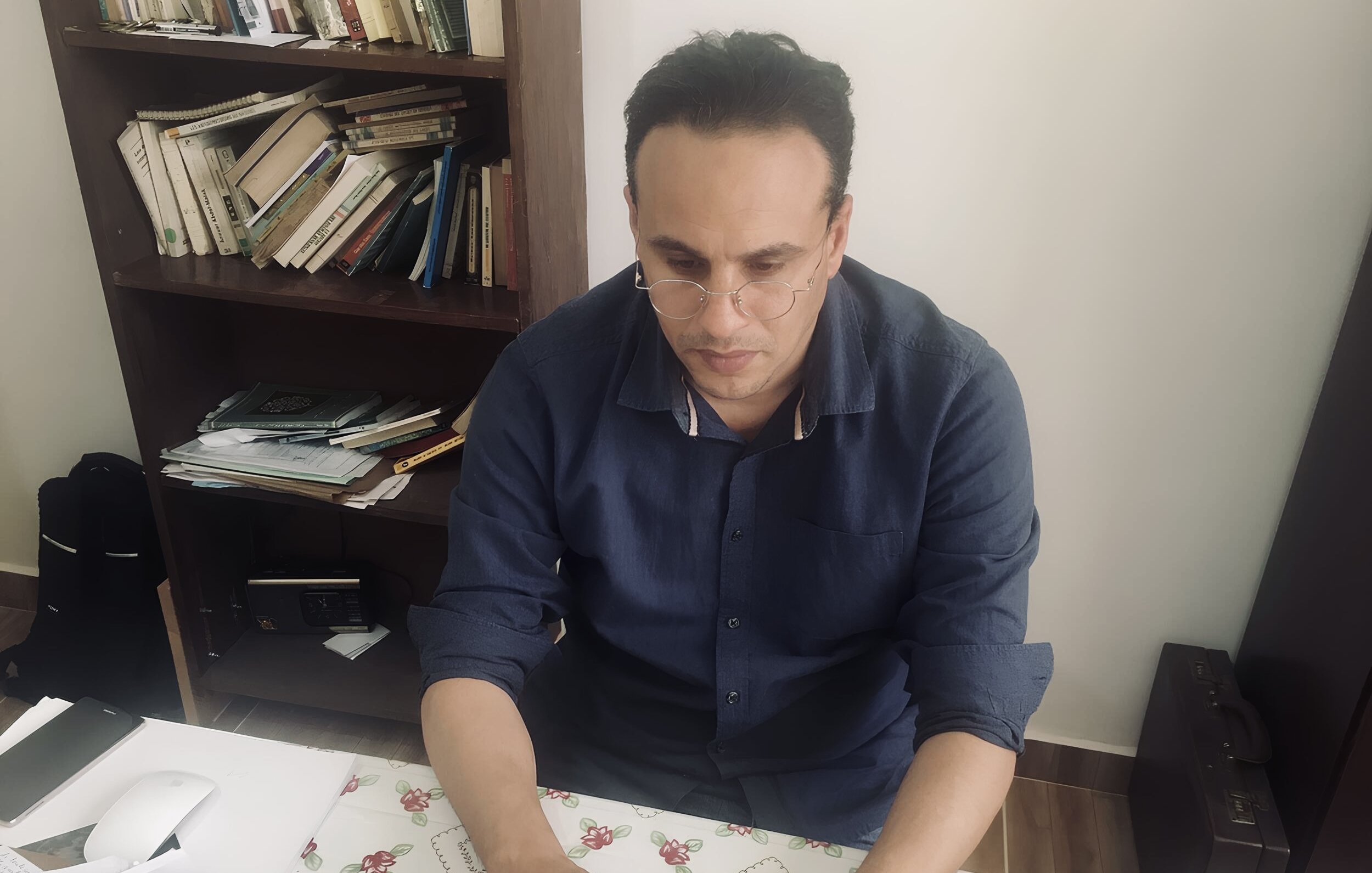 Nell'immagine si vede un uomo seduto a una scrivania. Indossa una camicia blu e occhiali. È concentrato mentre lavora su un computer portatile. La scrivania è disordinata, con fogli, un mouse e altri oggetti sparsi. Sullo sfondo si intravede una libreria con libri. L'ambiente sembra luminoso e semplice.