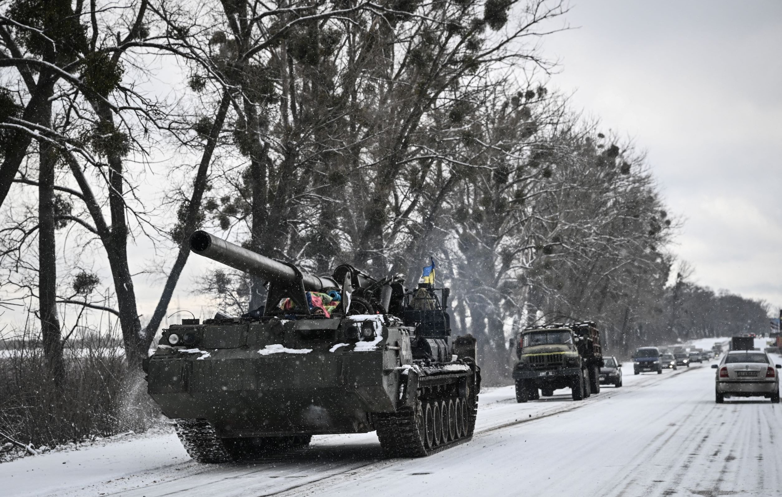 L'image montre un paysage hivernal avec de la neige recouvrant le sol. Un char militaire, probablement un tank, avance sur une route étroite bordée d'arbres. Des véhicules militaires sont également visibles en arrière-plan, créant une impression de mouvement dans un environnement froid et désert. L'atmosphère est marquée par des nuances de gris et de blanc, typiques d'un paysage d'hiver.