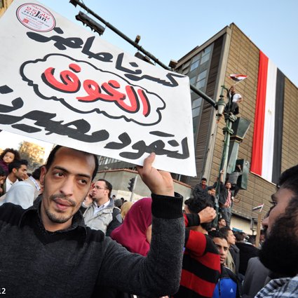 The image depicts a crowd gathered in an urban setting, likely during a protest or demonstration. In the foreground, a man holds a large sign with Arabic text, which translates to "Our demands are not to be ignored." In the background, there is a building displaying the Egyptian flag, suggesting a context related to political or social issues in Egypt. The scene conveys a sense of activism and public expression, with people rallying for their rights or changes in governance.