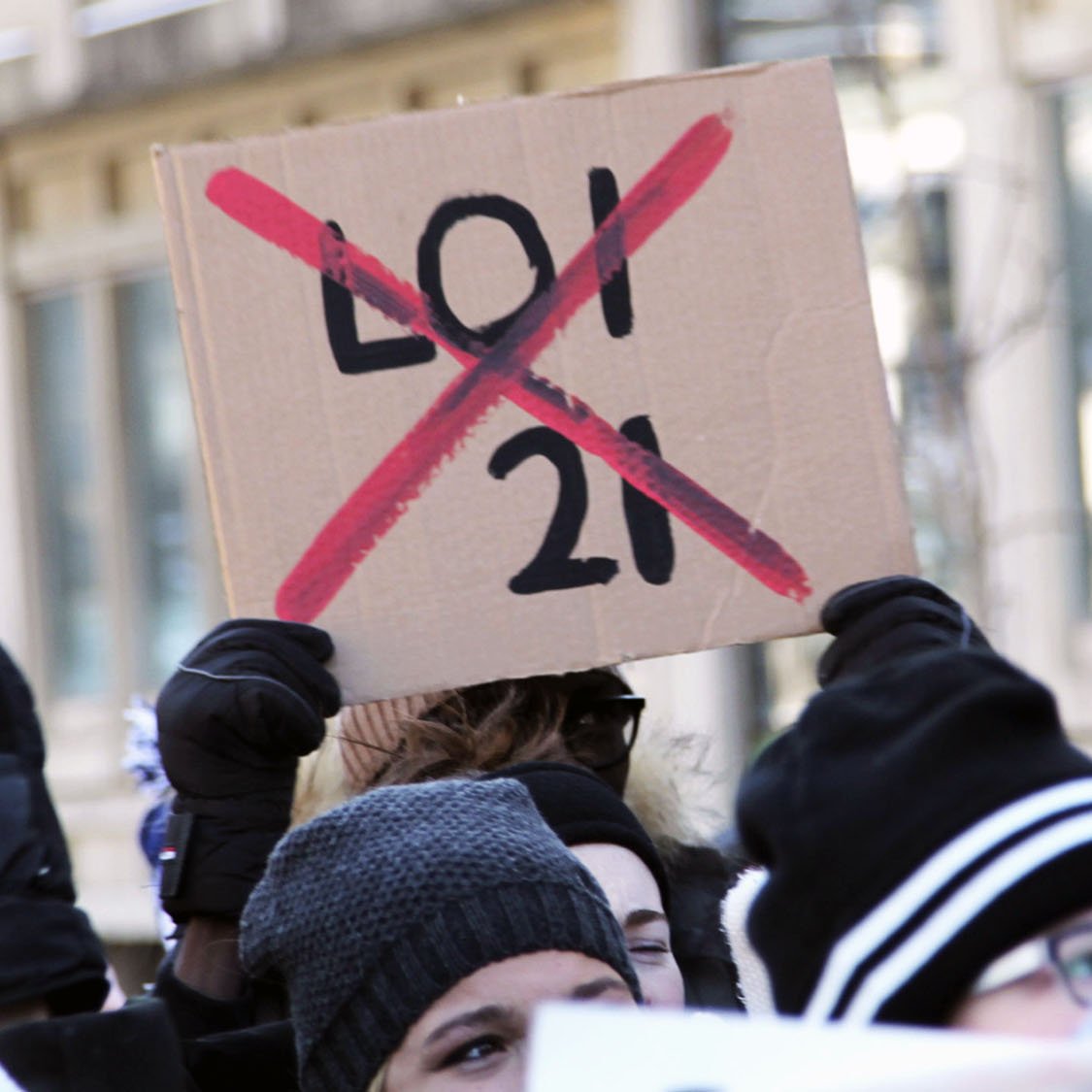 La imagen muestra a un grupo de personas participando en una manifestación. Algunas de ellas sostienen carteles, y uno de los más destacados es un cartel de cartón que dice "X" sobre el número "21", lo que indica oposición a esa ley o número específico. La multitud parece estar animada y comprometida con la causa que defienden. Las personas están abrigadas, usando gorros y guantes, lo que sugiere que hace frío.