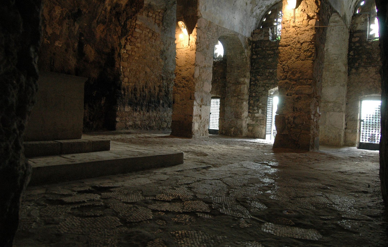The image depicts an interior space that appears to be a stone chamber or crypt. The walls are made of rough stone, showcasing an ancient architectural style. Light filters in through arched windows, casting a soft glow on the uneven stone floor. There are several columns supporting the ceiling, and a platform or altar can be seen against one wall. The atmosphere is quiet and austere, suggesting a place of historical or spiritual significance.