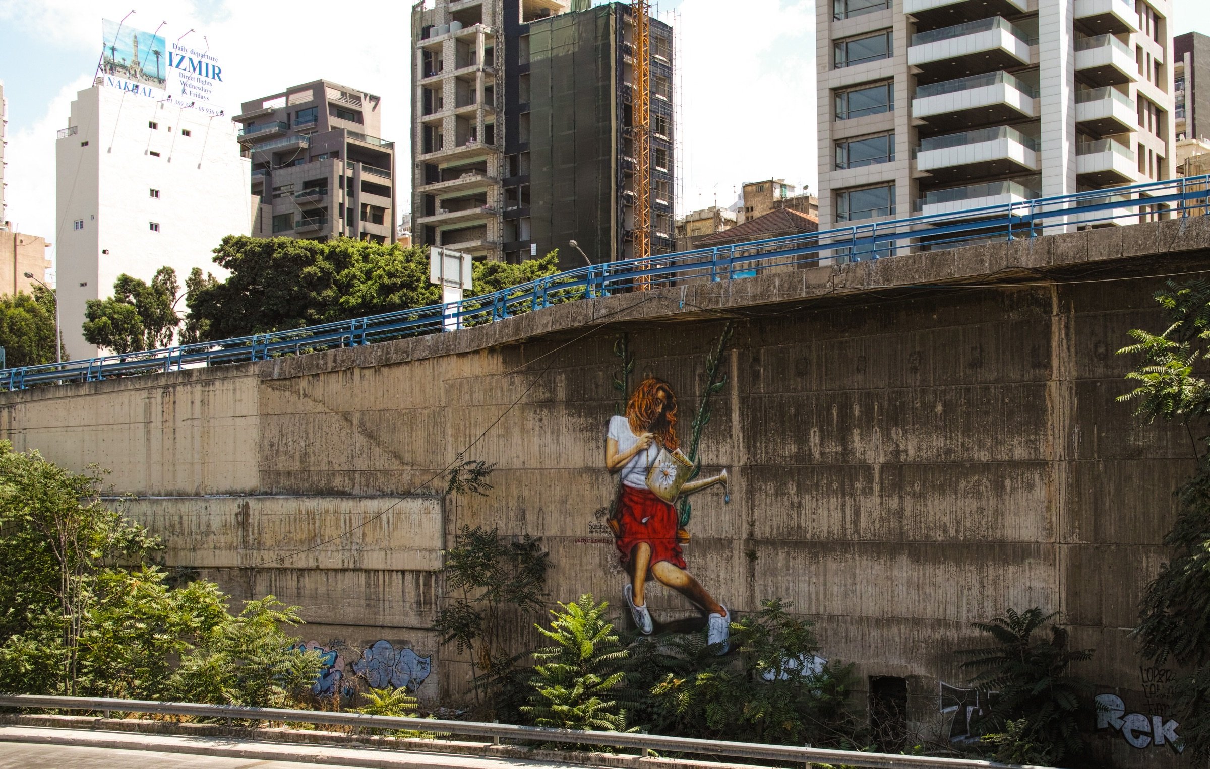 L'image montre un mur urbain en béton, partiellement recouvert de végétation. À droite, une grande peinture murale représente une personne en train de jouer à la balançoire, accrochée au mur. En arrière-plan, on aperçoit des bâtiments modernes, certains en construction, ce qui donne une ambiance de ville dynamique. La scène mélange l'art urbain avec des éléments naturels, créant un contraste intéressant entre le béton et la verdure.
