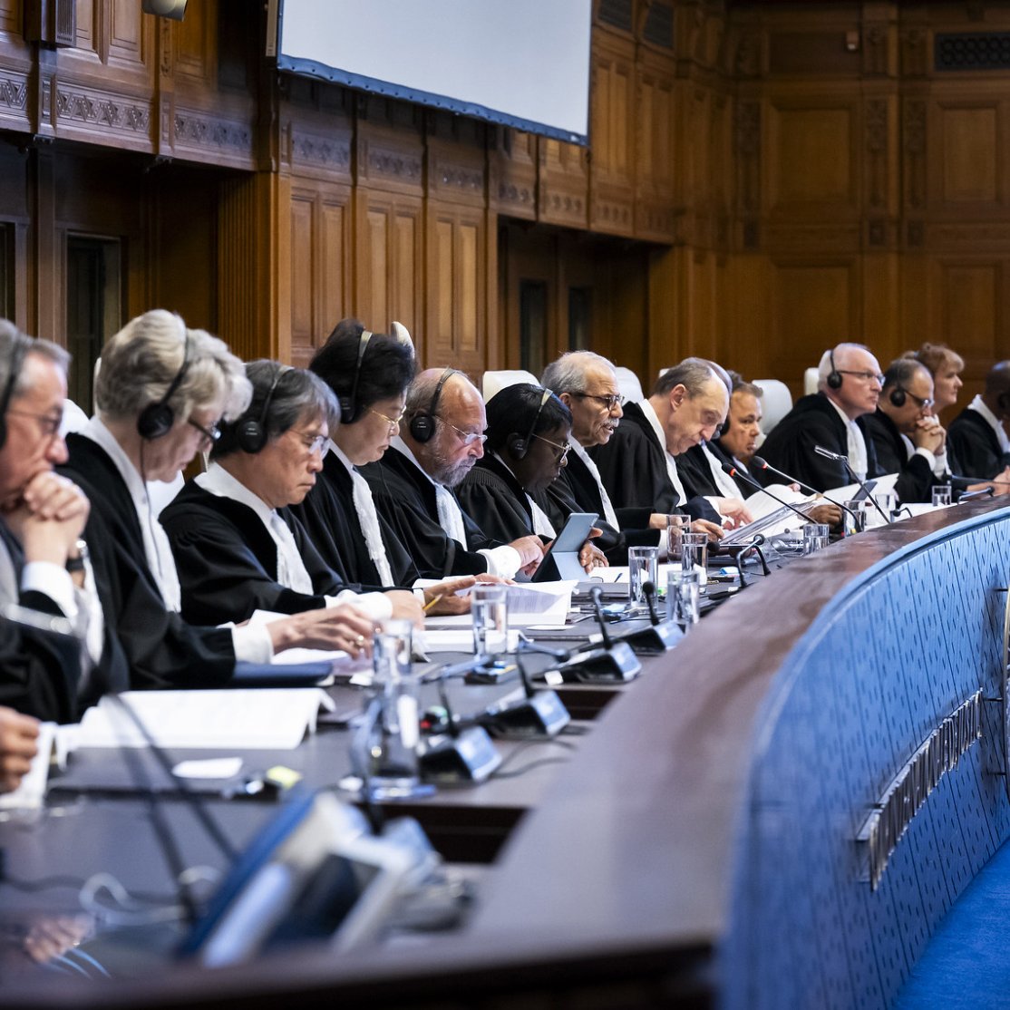 L'image montre une salle de tribunal où plusieurs juges sont assis en ligne dans un espace formel et bien éclairé. Ils portent des robes noires et des écouteurs, symbolisant une séance judiciaire importante. Le décor est élégant, avec des murs en bois et un grand écran au fond. Les juges semblent concentrés sur les documents devant eux, participant à une discussion ou à une audience. L'atmosphère est sérieuse, en accord avec la nature de leurs fonctions.