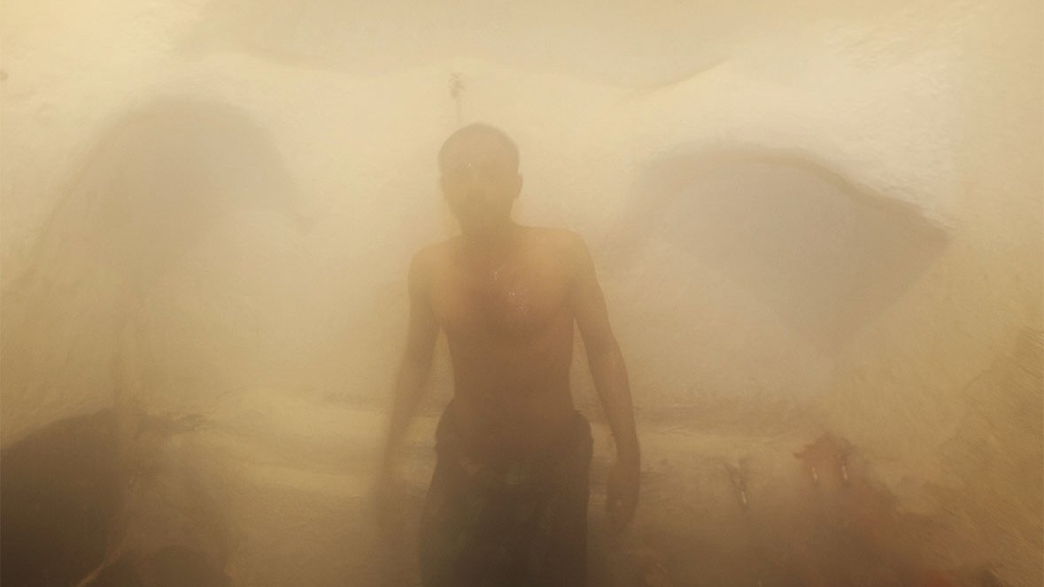 L'image montre un homme dans un environnement humide et brumeux, probablement à l'intérieur d'un hammam ou d'un sauna. La vapeur crée une atmosphère indistincte et floue, rendant les contours des objets difficilement visibles. L'homme est torse nu et semble se tenir debout, enveloppé par la vapeur qui l'entoure, ajoutant une sensation de chaleur et de sérénité à la scène. Les murs sont d'apparence texturée, typiques des espaces de bien-être.