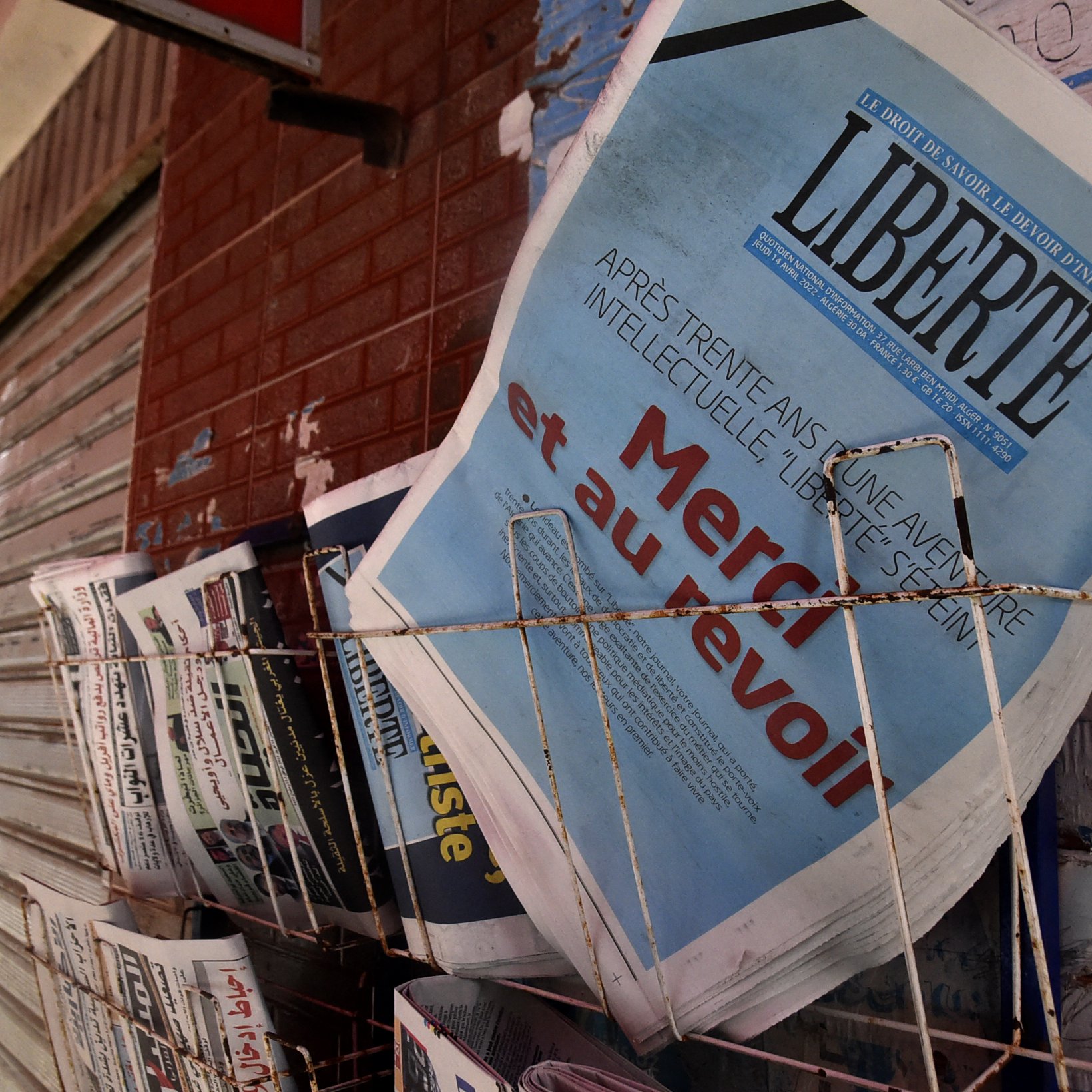 L'image montre un présentoir de journaux, où plusieurs titres sont visibles. Le journal en premier plan affiche le mot "LIBERTÉ" en lettres majuscules, accompagnés d'un sous-titre qui semble évoquer un sujet d'actualité. Les autres journaux à côté sont disposés de manière ordonnée, avec des titres variés, ce qui suggère un point de vente de presse dans un environnement urbain. Le mur en arrière-plan, ainsi que le toît rouge au-dessus, ajoutent à l'atmosphère locale de la scène.