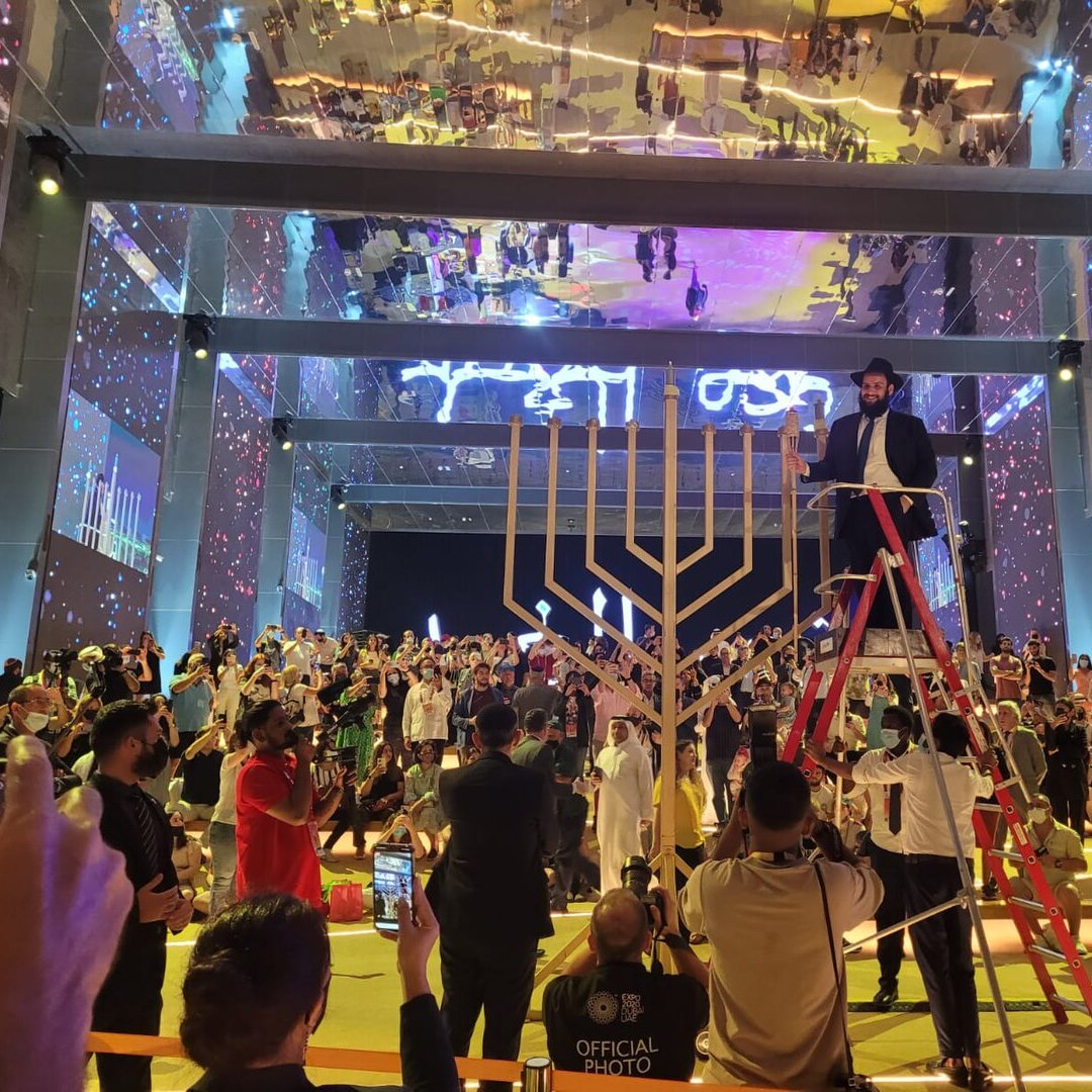 L'immagine mostra un evento festivo e affollato in un grande spazio, con una decorazione vibrante e luci colorate. Al centro, c'è una grande menorah, simbolo della tradizione ebraica, e una persona in cima a una scala che sembra essere coinvolta nella cerimonia di accensione. Il pubblico è composto da diverse persone in abiti tradizionali e moderni, tutte rivolte verso il palco. Il soffitto è riflettente, amplificando l'atmosfera festiva, mentre sullo sfondo si possono notare schermi luminosi e decorazioni artistiche. L'intera scena trasmette un senso di celebrazione e comunità.