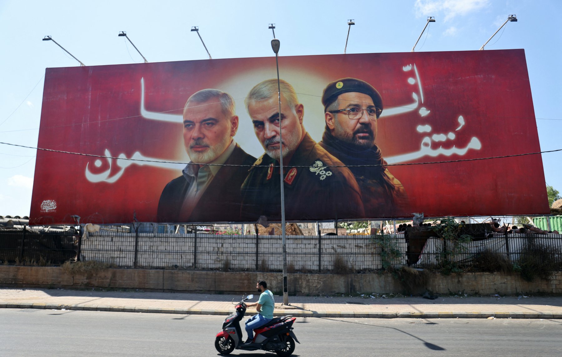 الصورة تظهر لوحة إعلانات ضخمة تحمل صور ثلاثة أشخاص. الخلفية حمراء اللون، وتبدو وكأنها تتضمن رسائل وطنية أو سياسية. في المقدمة، يوجد رجلان يواجهان الكاميرا ورجل آخر يُظهر جزءًا من وجهه، مما يوحي بأهمية شخصياتهم في السياق المعروض. في أسفل اللوحة، يُظهر شخص يقود دراجة نارية، مما يضيف حركة في الصورة ويعكس الحياة اليومية في المدينة.