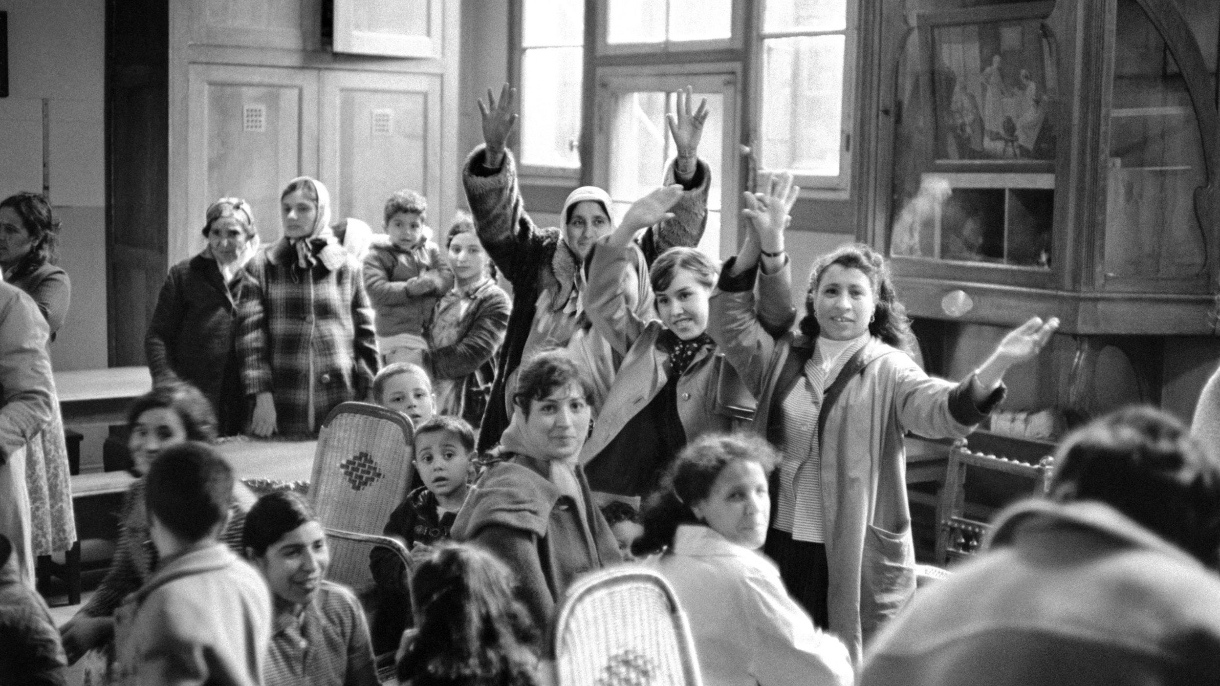 L'image en noir et blanc montre un groupe de personnes dans une pièce, probablement un lieu communautaire. Plusieurs adultes et enfants sont présents, certains levant les bras en signe d'encouragement ou d'émerveillement. L'atmosphère semble conviviale, avec des visages qui expriment de la joie ou de l'excitation. On peut voir des fauteuils en osier et un grand meuble en bois à l'arrière-plan, ajoutant une touche de chaleur à l'environnement. La lumière naturelle entre par de grandes fenêtres, illuminant la scène.