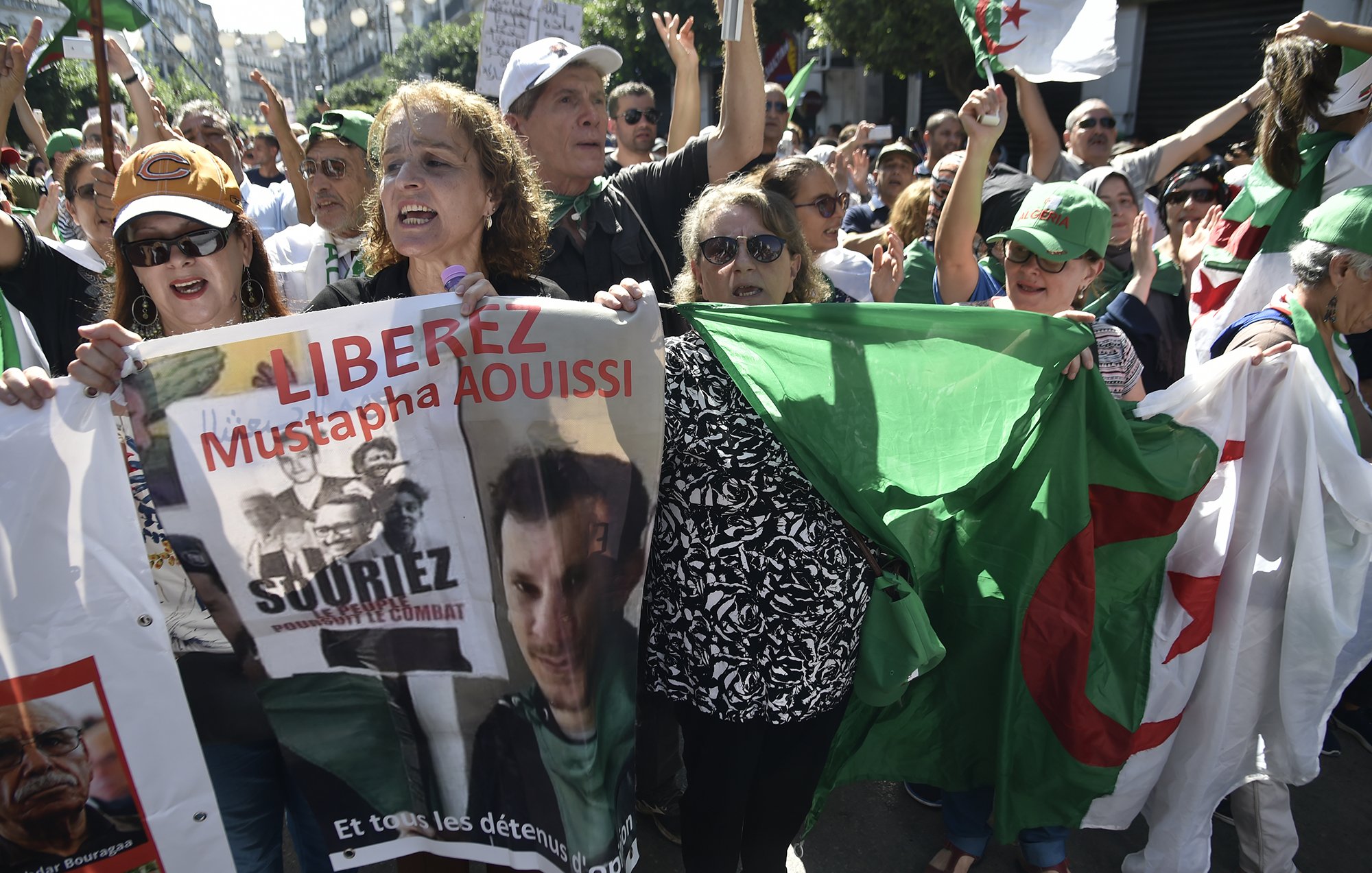 الصورة تظهر مجموعة من المتظاهرين في الجزائر، يحملون لافتات وأعلام. يبدو أن هناك نساء ورجال في المقدمة، ويعبرون عن مطالبات سياسية. إحدى اللافتات تحمل اسم "مصطفى عويصي" وعبارة "حرروا". الأجواء توحي بالحماسة والمطالبة بالتغيير.