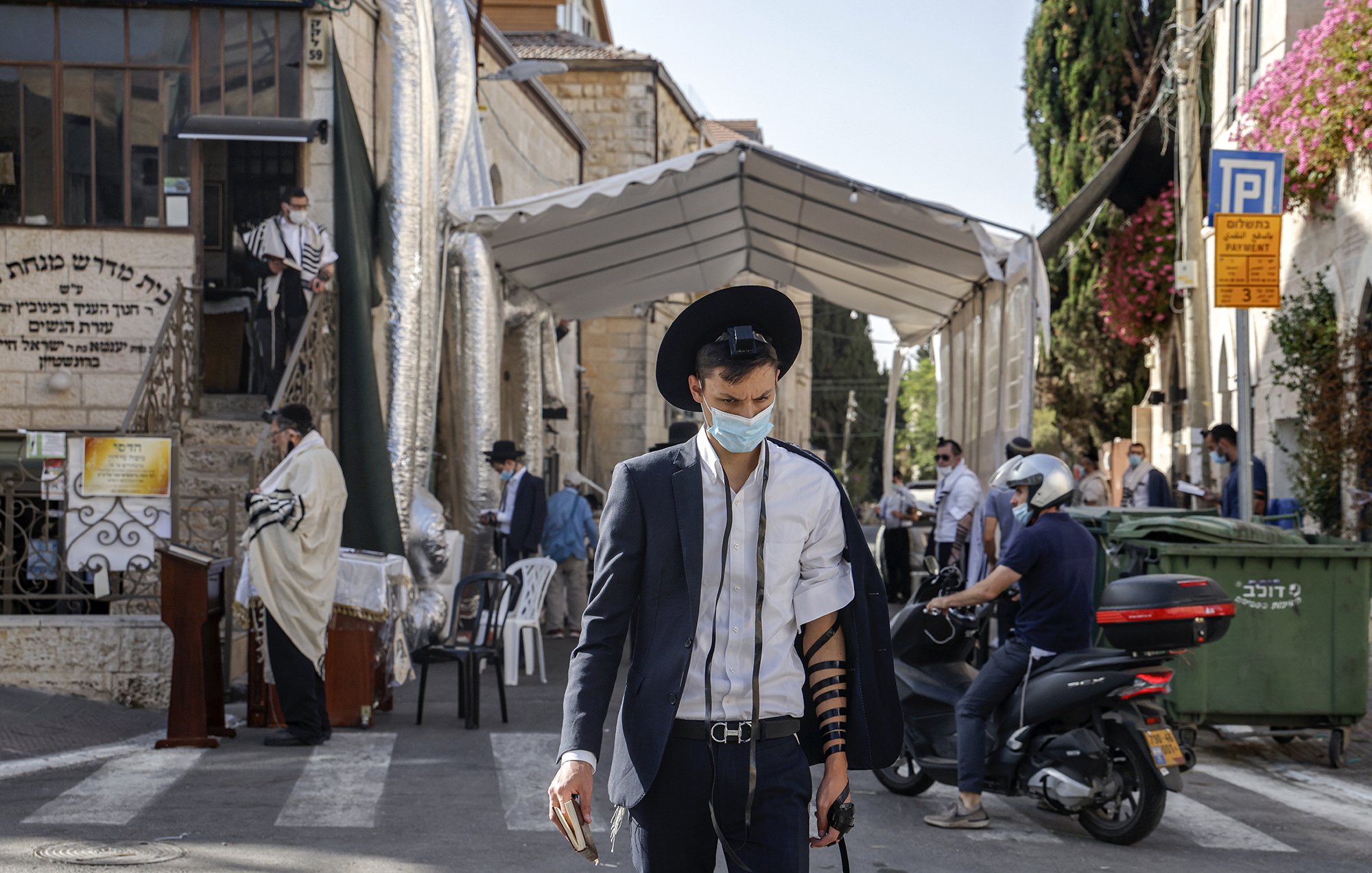 The image shows a street scene in an urban area, likely in a Jewish neighborhood. A young man wearing a mask and traditional attire, including a black hat and a suit, is walking while wearing tefillin (phylacteries) on his arm. In the background, there is a large tent set up for what appears to be a religious gathering, with several other individuals, some dressed in traditional clothing, visible. The setting is sunny, and there are various buildings, including signs and greenery, contributing to the atmosphere of the scene.