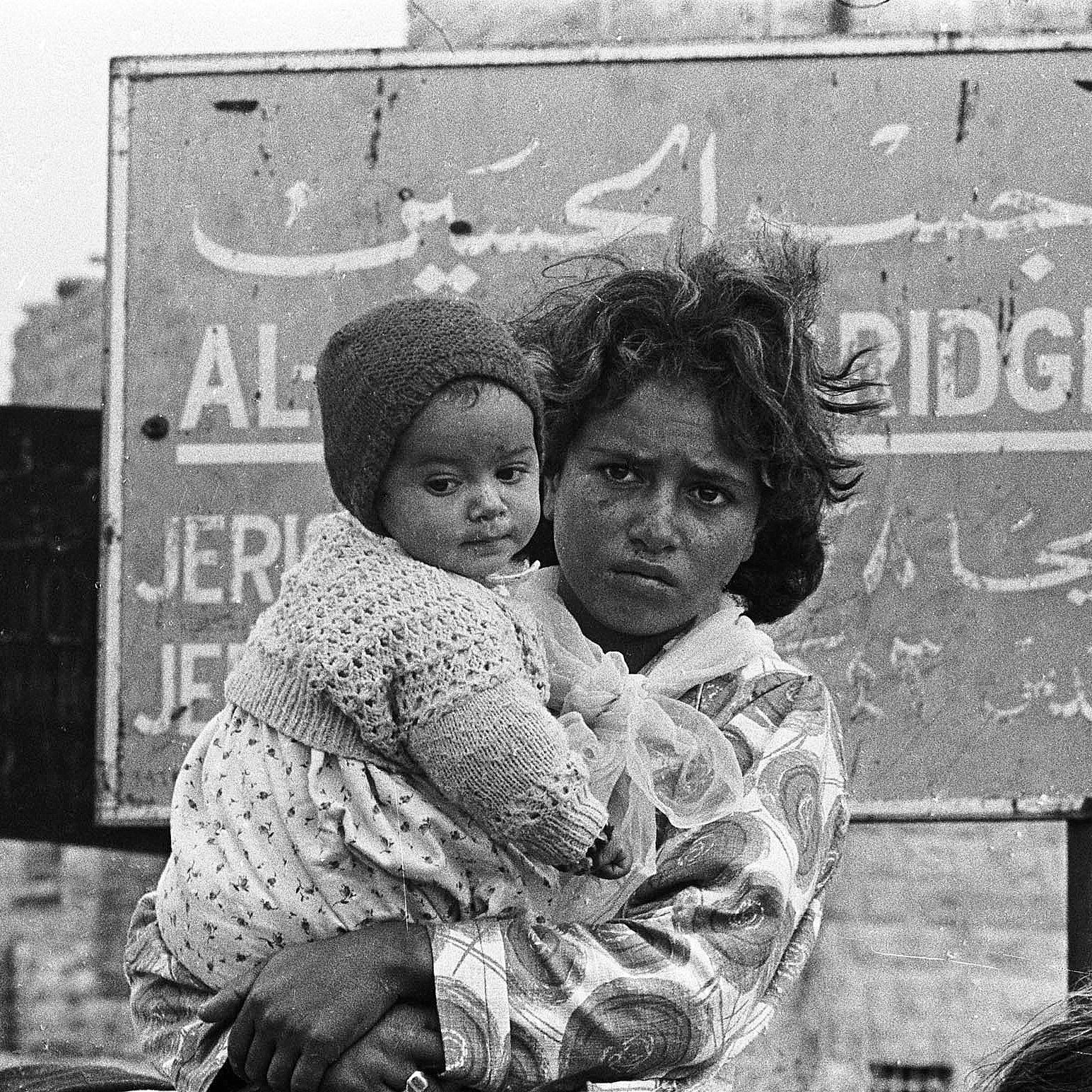 The image depicts a young woman holding a baby, both appearing to be in a challenging environment. The woman has a serious expression on her face, and her hair is unkempt. The baby, swaddled in a heavy garment, is held close to her. In the background, there's a sign written in Arabic, which seems to indicate a location, possibly related to a city or area. The overall atmosphere conveys a sense of hardship and resilience. The image is in black and white, emphasizing the emotional weight of the scene.