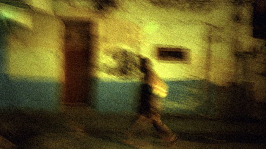 L'image présente une scène nocturne floue, où une silhouette se déplace le long d'un mur aux teintes décolorées, combinant des nuances de vert et de bleu. La texture du mur semble usée et vieillie, suggérant un environnement urbain. La personne, portant un sac, marche rapidement, créant un effet de mouvement qui renforce l'impression de dynamisme et d'évasion dans cette ambiance mystérieuse.