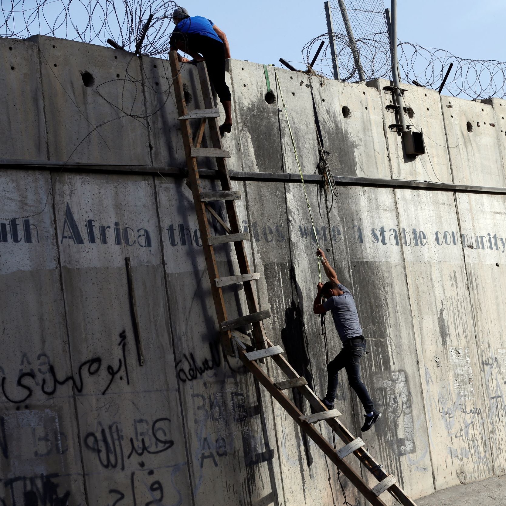 La imagen muestra a dos personas subiendo una pared de concreto alta. Una de ellas está en la parte superior, mientras que la otra está en una escalera de madera, intentando escalar. En la pared hay alambres de púas en la parte superior y también se pueden ver algunas inscripciones y grafitis en varios idiomas. El ambiente parece ser tenso y el contexto sugiere una situación de conflicto o segregación.