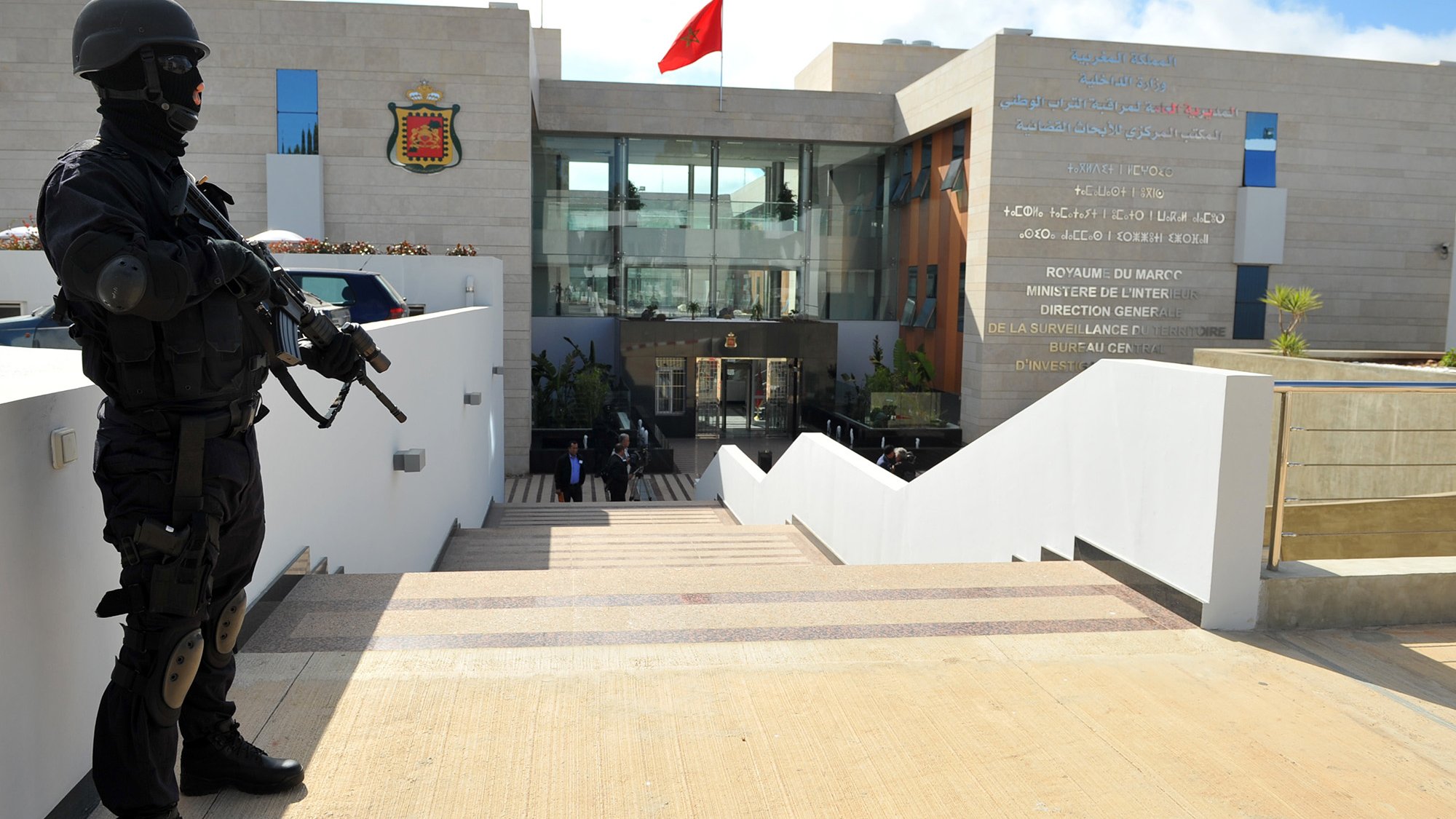 L'image montre un bâtiment moderne et officiel, probablement un ministère ou une institution gouvernementale, orné d'un drapeau marocain. À l'avant, un soldat en tenue noire, portant un masque et une arme, se tient en position de garde sur des escaliers menant à l'entrée du bâtiment. Des inscriptions en français et en arabe sont visibles sur le mur à côté de l'entrée, indiquant le nom de l'institution. L'environnement semble sécurisé et bien entretenu, avec quelques plantes décoratives.
