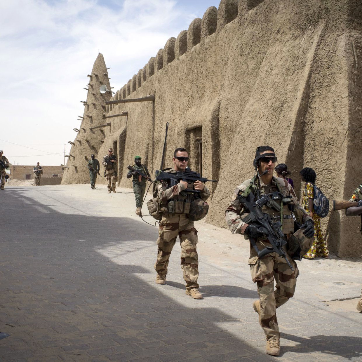 La imagen muestra a un grupo de soldados que caminan por una calle en una zona con arquitectura de barro. A su alrededor, se pueden ver estructuras típicas del lugar, como muros de adobe. Los soldados llevan equipos militares y armamento, y algunos parecen estar en formación o realizando patrullas. La escena sugiere un ambiente de seguridad o una operación militar en un contexto urbano o semiurbano.
