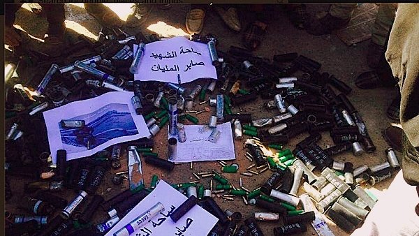 L'image montre un grand nombre de cartouches de munitions et de projectiles disposés au sol. Certaines de ces munitions sont accompagnées de panneaux ou de feuilles de papier avec des inscriptions en arabe. On peut voir des personnes autour de cette disposition, mais leurs visages ne sont pas visibles. L'ensemble de la scène semble évoquer une situation liée à la confiscation ou à la destruction d'armements.