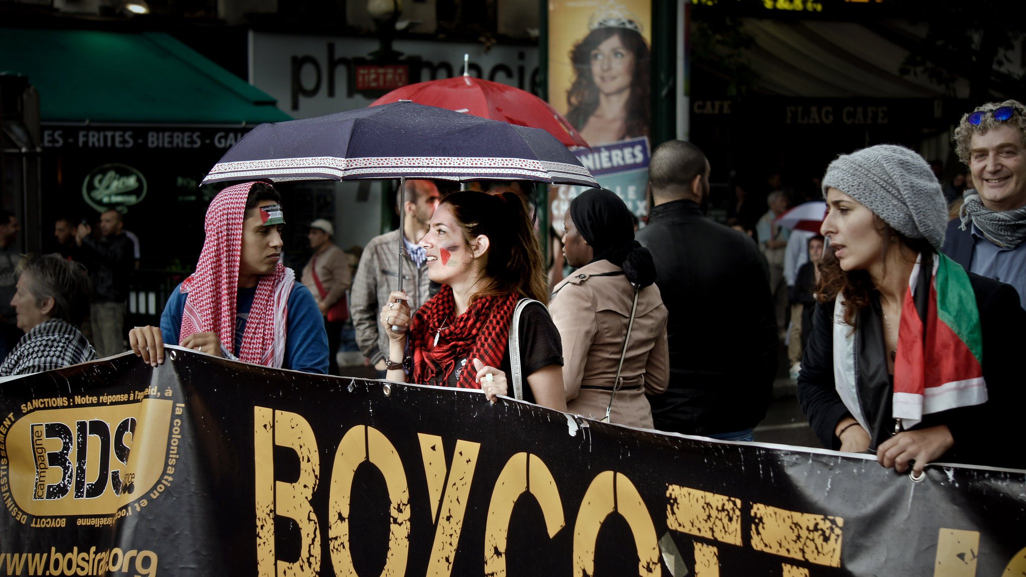 L'image montre un groupe de personnes participant à une manifestation. Au premier plan, on voit une femme tenant un grand panneau qui dit "BOYCOTT". Elle est entourée de plusieurs manifestants, dont certains portent des accessoires ou des vêtements aux couleurs de différents pays. Il y a aussi des parapluies, probablement à cause de la météo. L'ambiance semble engagée, avec un mélange de détermination et de solidarité parmi les participants.