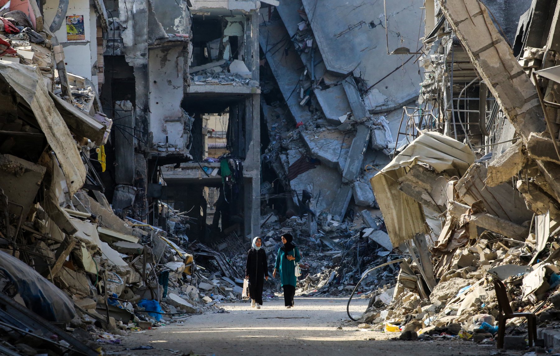 L'image montre un paysage de destruction urbaine, avec des bâtiments effondrés sur les côtés d'une rue. On peut apercevoir deux personnes marchant au milieu des décombres. L'atmosphère est sombre et désolée, avec des ruines qui témoignent d'une catastrophe récente. Les déchets et les restes de matériaux de construction jonchent le sol, créant un sentiment de désolation.