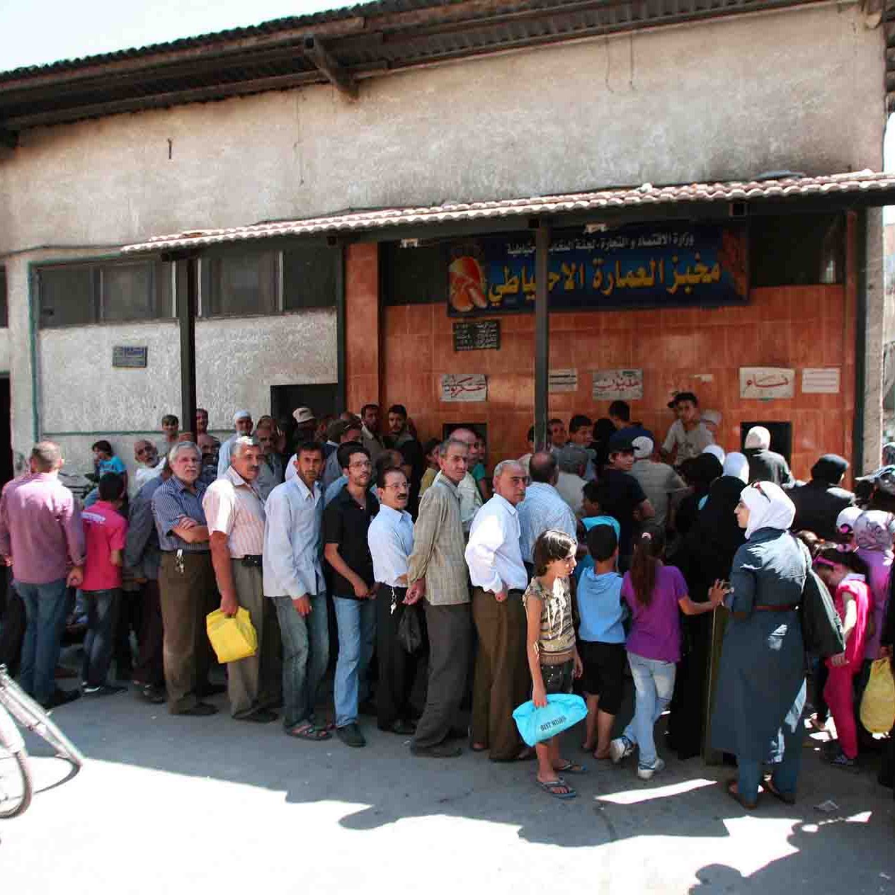 L'immagine mostra una lunga fila di persone che si trovano all'esterno di un edificio. La scena sembra essere in un contesto urbano, probabilmente in una città del Medio Oriente. Le persone nella fila sono di diverse età e indossano abiti vari, alcuni dei quali tradizionali. Alcuni individui portano sacche o borse, e si può notare un'atmosfera di attesa e di comunità. Sullo sfondo, ci sono edifici semplici con elementi architettonici tipici della regione.