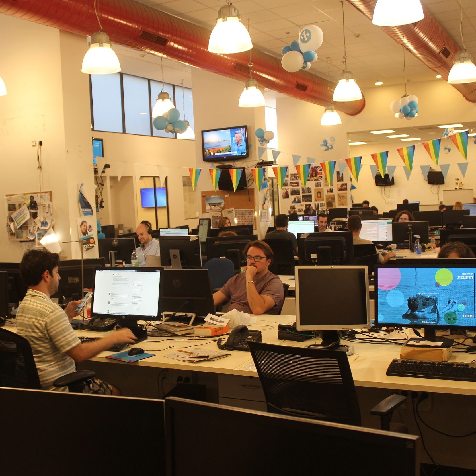L'immagine mostra un ambiente di lavoro in un ufficio moderno. Si possono vedere diverse scrivanie con computer e monitor. Alcune persone sono immerse nel loro lavoro, mentre altre interagiscono tra di loro. L'ufficio è decorato con palloncini blu e festoni colorati, creando un'atmosfera vivace e informale. Le pareti sono adornate con fotografie e materiali di lavoro, e la luce naturale entra attraverso le grandi finestre.