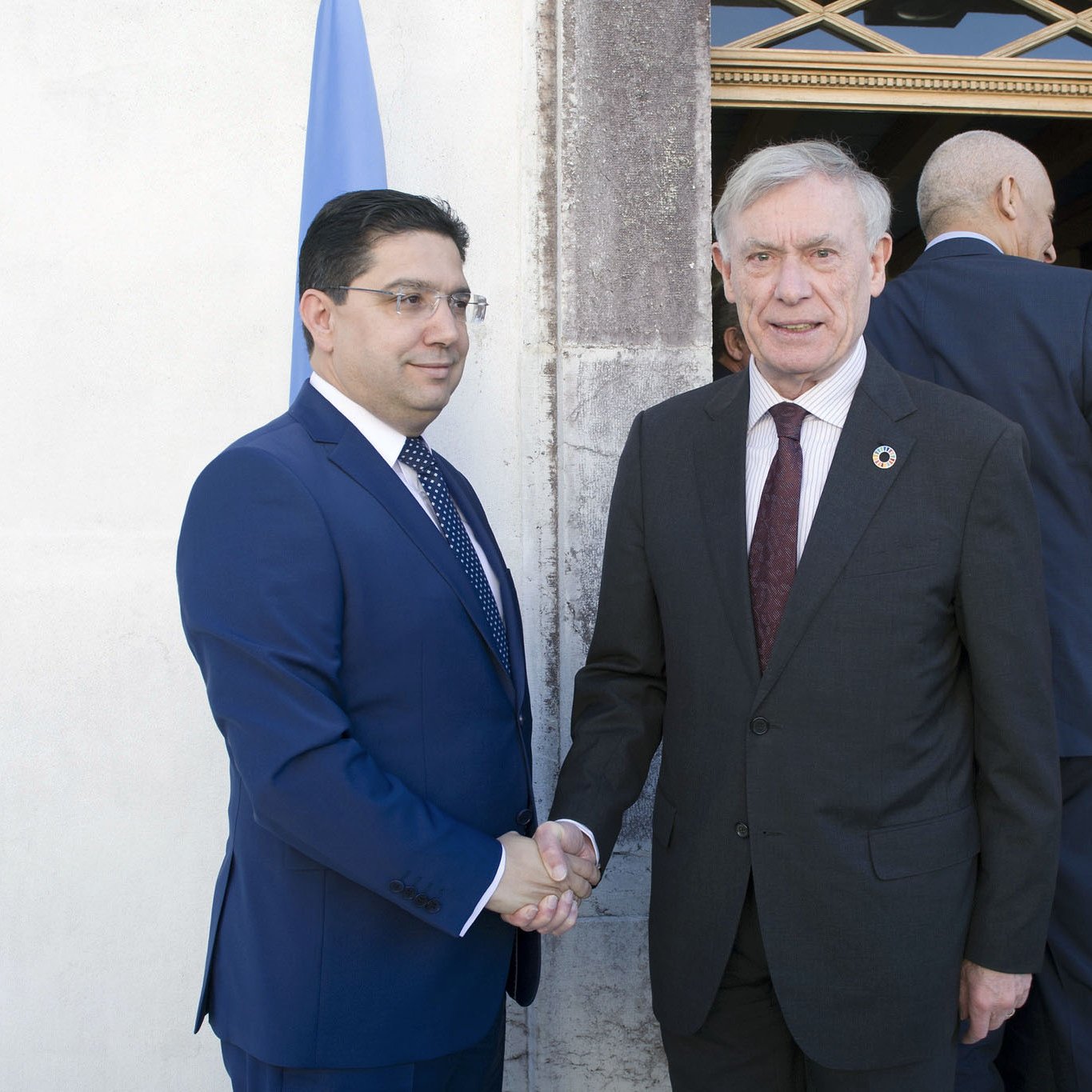 En la imagen se puede ver a dos hombres en un ambiente formal. Uno de ellos está vestido con un traje oscuro y una corbata, mientras que el otro lleva un traje azul. Ambos están dándose la mano, lo que sugiere un saludo o un acuerdo. En el fondo hay una puerta y una bandera, lo que indica que están en un entorno institucional o diplomático. La escena transmite un sentido de cordialidad y oficialidad.