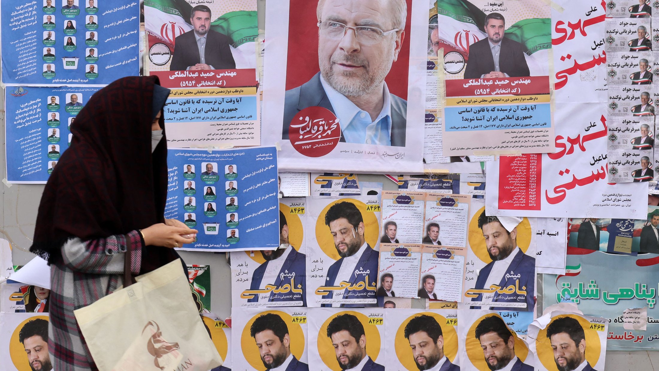 L'image montre un mur recouvert de diverses affiches électorales. On peut voir plusieurs portraits de candidats, accompagnés de slogans et d'informations. Au premier plan, une femme portant un hijab et tenant un sac semble marcher devant ces affiches, ce qui suggère une ambiance électorale. Les couleurs des affiches sont vives et variées, ajoutant à l'aspect animé de la scène.