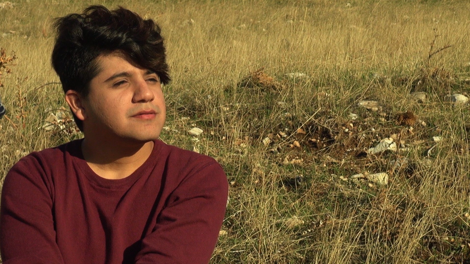 L'image montre une personne assise dans un champ de herbes hautes. La personne a les cheveux foncés et bouclés, et elle semble pensive, regardant au loin. Elle porte un pull de couleur bordeaux. L'environnement est naturel, avec une lumière douce, ce qui crée une atmosphère tranquille.