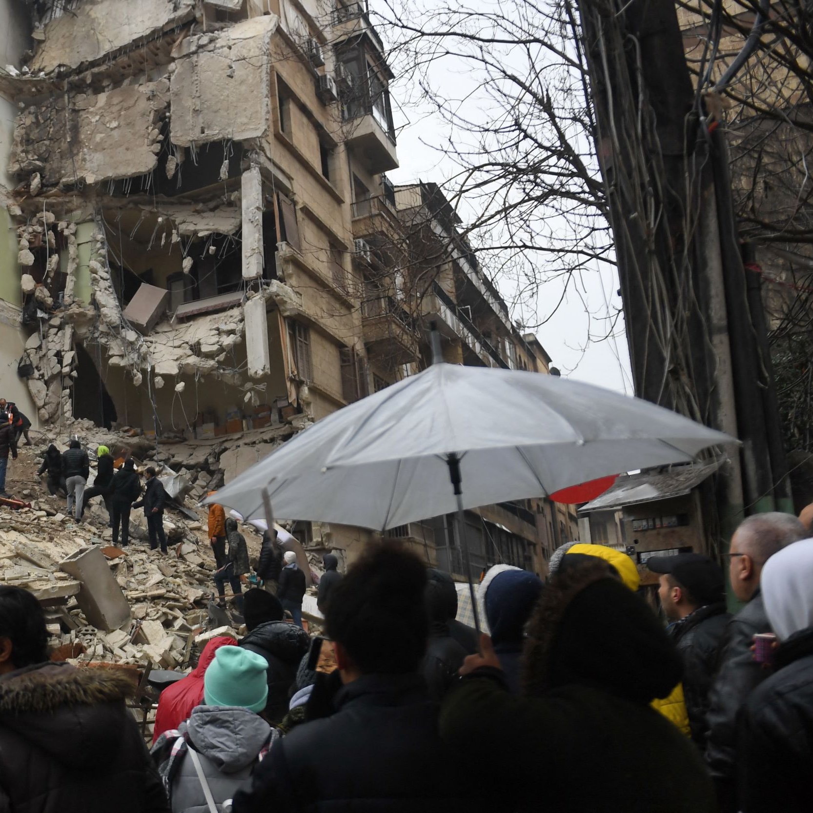 این تصویر یک صحنه ویران شده از یک زمین‌لرزه را نمایش می‌دهد. در تصویر، تعدادی از افرادی که به سمت محل خرابی رفته‌اند، دیده می‌شوند. ساختمان‌ها به شدت آسیب دیده و برخی از دیوارها و تکیه‌گاه‌ها فرو ریخته‌اند. همچنین چند نفر با چتر در حال نظارت و کمک به دیگران هستند. مردم با نگران و متعجب به این صحنه نگاه می‌کنند، و احساس غم و ناراحتی در فضا حاکم است.