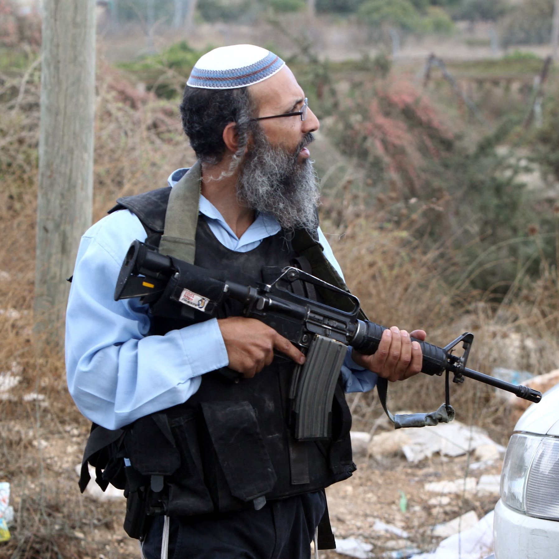 L'image montre un homme qui porte une kippa et une veste de protection. Il est armé d'un fusil automatique et se tient près d'une voiture dans un environnement extérieur, avec de la végétation et des débris autour de lui. Son expression et sa posture suggèrent qu'il est en alerte ou surveille les alentours.