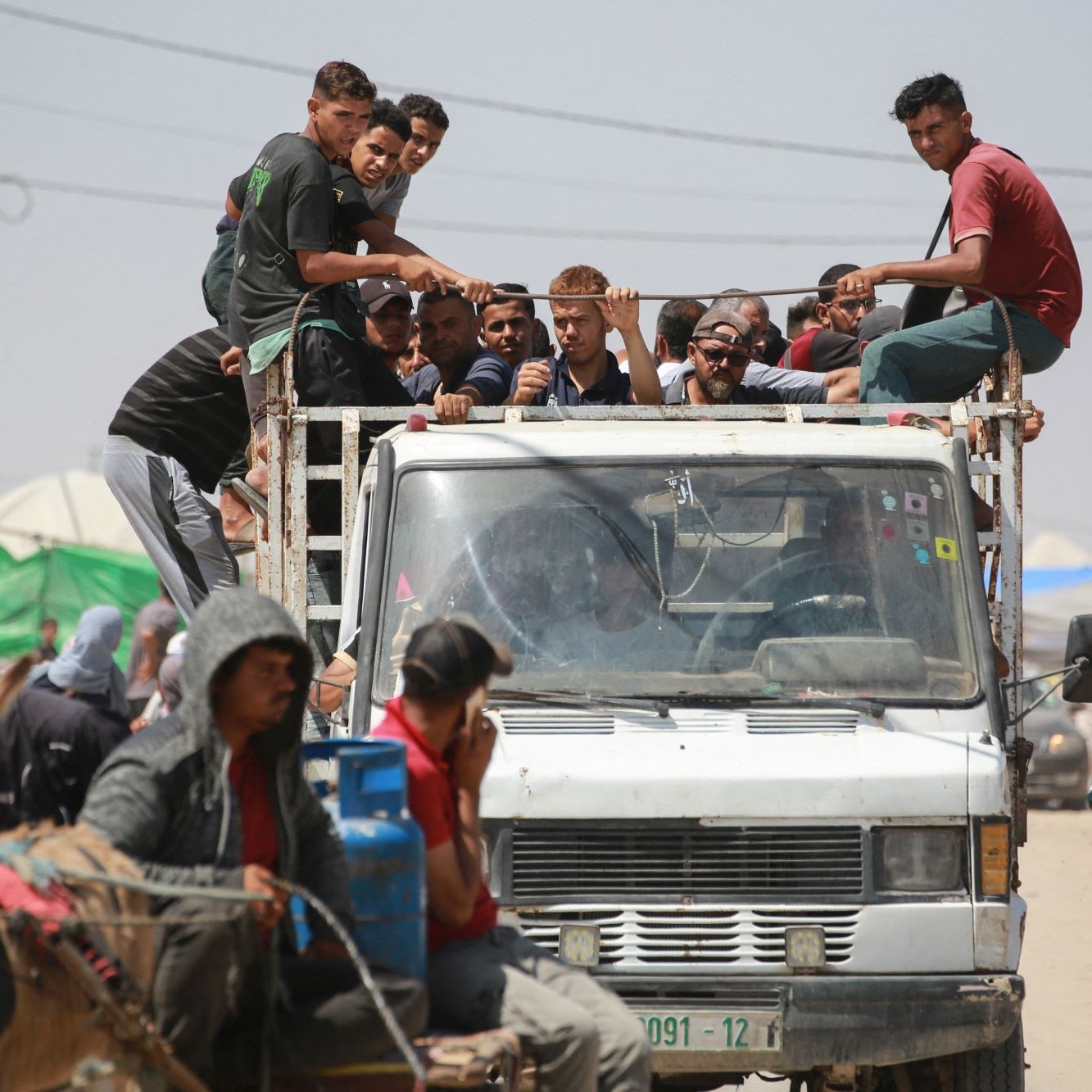 L'immagine mostra un gruppo di persone che si trovano su un camion, alcuni di loro sono seduti sul retro del veicolo, mentre altri sono in piedi. Sul lato destro ci sono persone su un carro trainato da un animale, che sembra essere un cavallo. In fondo si intravedono tende e strutture, suggerendo un contesto urbano o di accampamento. L'atmosfera sembra vivace, con diverse persone che interagiscono tra loro.