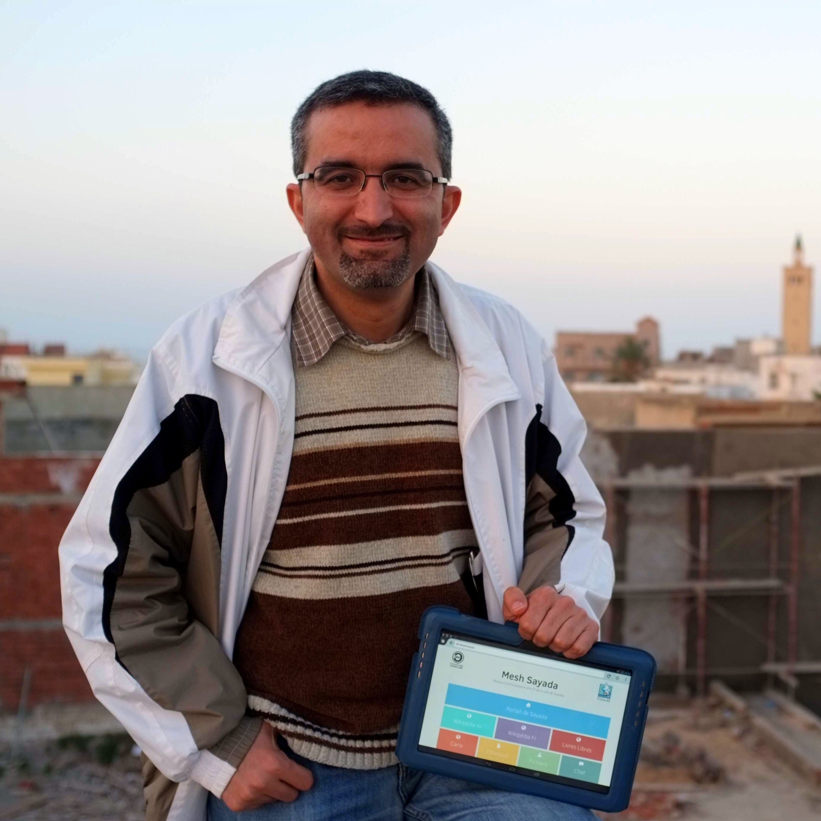 L'image montre un homme souriant debout sur un toit, tenant une tablette. Il porte une veste blanche et un pull rayé. En arrière-plan, on peut voir des bâtiments et un minaret au loin, indiquant un paysage urbain. L'éclairage suggère un moment de la fin de journée, avec un ciel aux teintes douces.