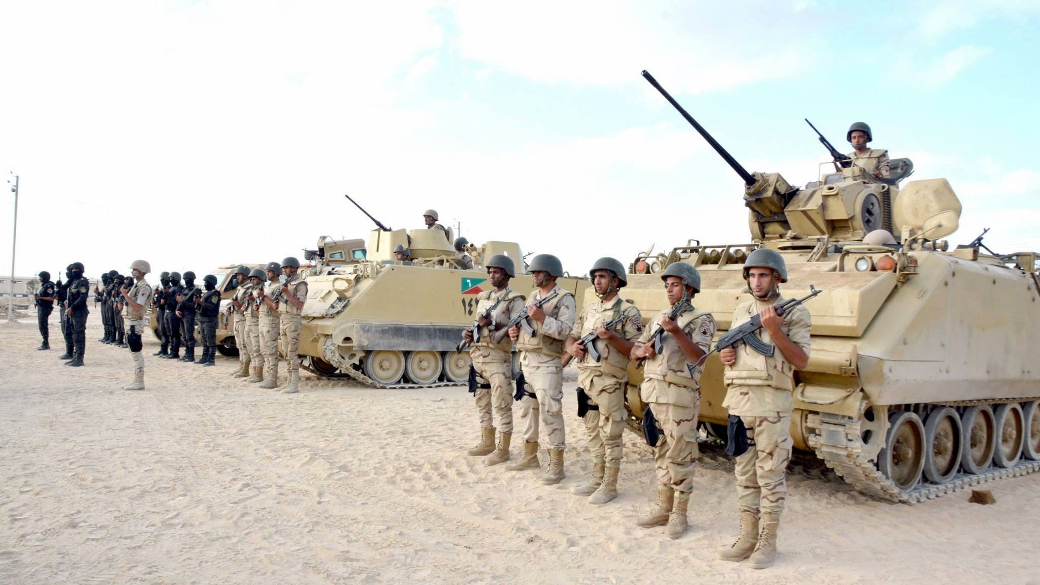 L'image montre une formation militaire dans un environnement désertique. Au premier plan, des soldats en uniforme, armés et en position de garde, se tiennent alignés. En arrière-plan, on aperçoit plusieurs véhicules blindés, dont certains sont équipés de tourelles. Le ciel est clair, et le sol est sablonneux, typique des zones désertiques. Les soldats semblent concentrés et prêts pour une mission.