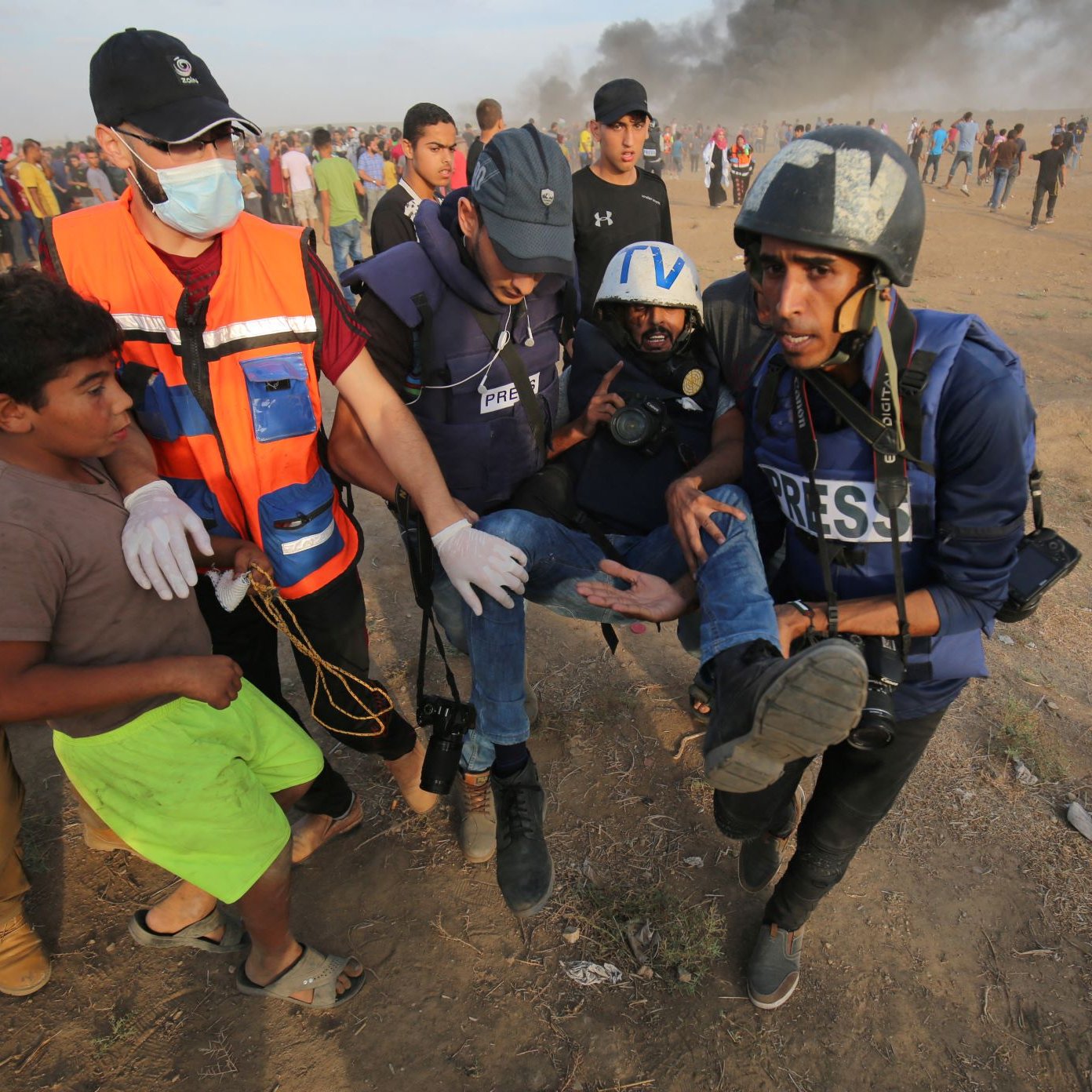 L'immagine mostra un gruppo di persone in una situazione di emergenza. Alcuni uomini, vestiti con giubbotti di protezione e tesserini "PRESS", stanno aiutando un uomo che è visibilmente ferito, portandolo via dal luogo del conflitto. Sullo sfondo si vede del fumo, suggerendo che ci sia stata un'esplosione o un incendio. Un ragazzo in primo piano osserva la scena con attenzione. L'atmosfera è tesa e drammatica, evidenziando una situazione di crisi.