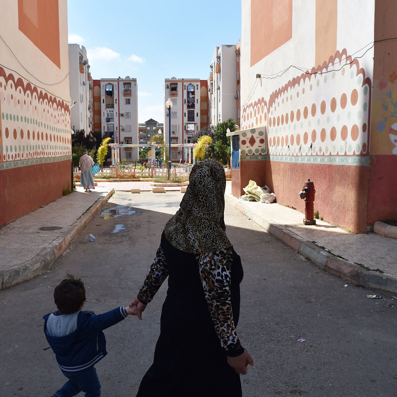 La imagen muestra una escena de una calle en un barrio residencial. En el primer plano, una mujer con un hiyab sostiene de la mano a un niño pequeño mientras caminan. Al fondo, se pueden ver edificios de apartamentos con pinturas decorativas en las paredes. El ambiente parece tranquilo y iluminado por la luz del día, y hay un pequeño espacio ajardinado en el centro, lo que sugiere un lugar comunitario.