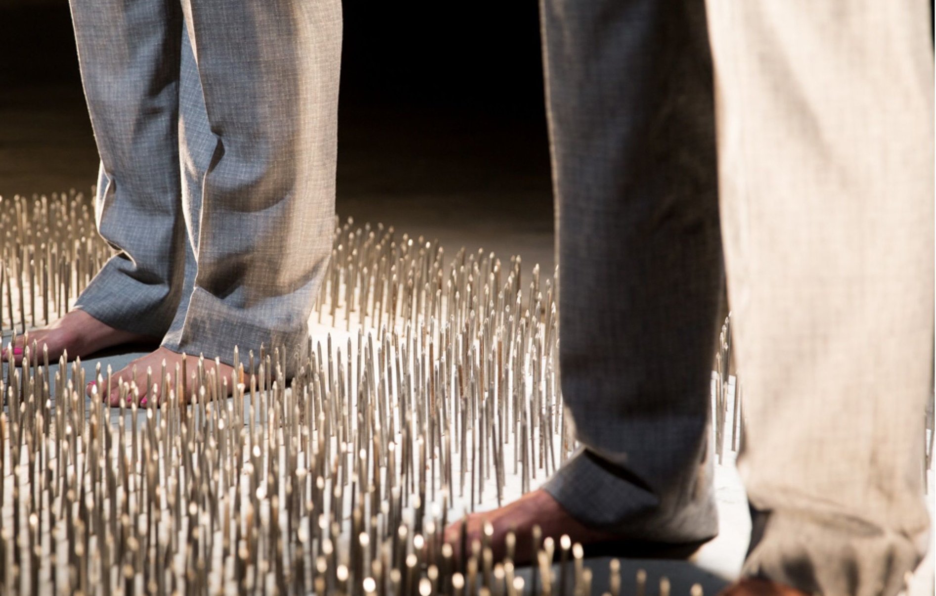 L'image montre deux personnes se tenant debout sur une surface couverte de nombreux picots métalliques. Les individus portent des pantalons clairs, et leurs pieds nus touchent les picots. La scène semble mettre en avant un contraste fort entre le confort des vêtements et l'inconfort potentiel de la surface sur laquelle ils se tiennent, créant une atmosphère à la fois intrigante et dérangeante.